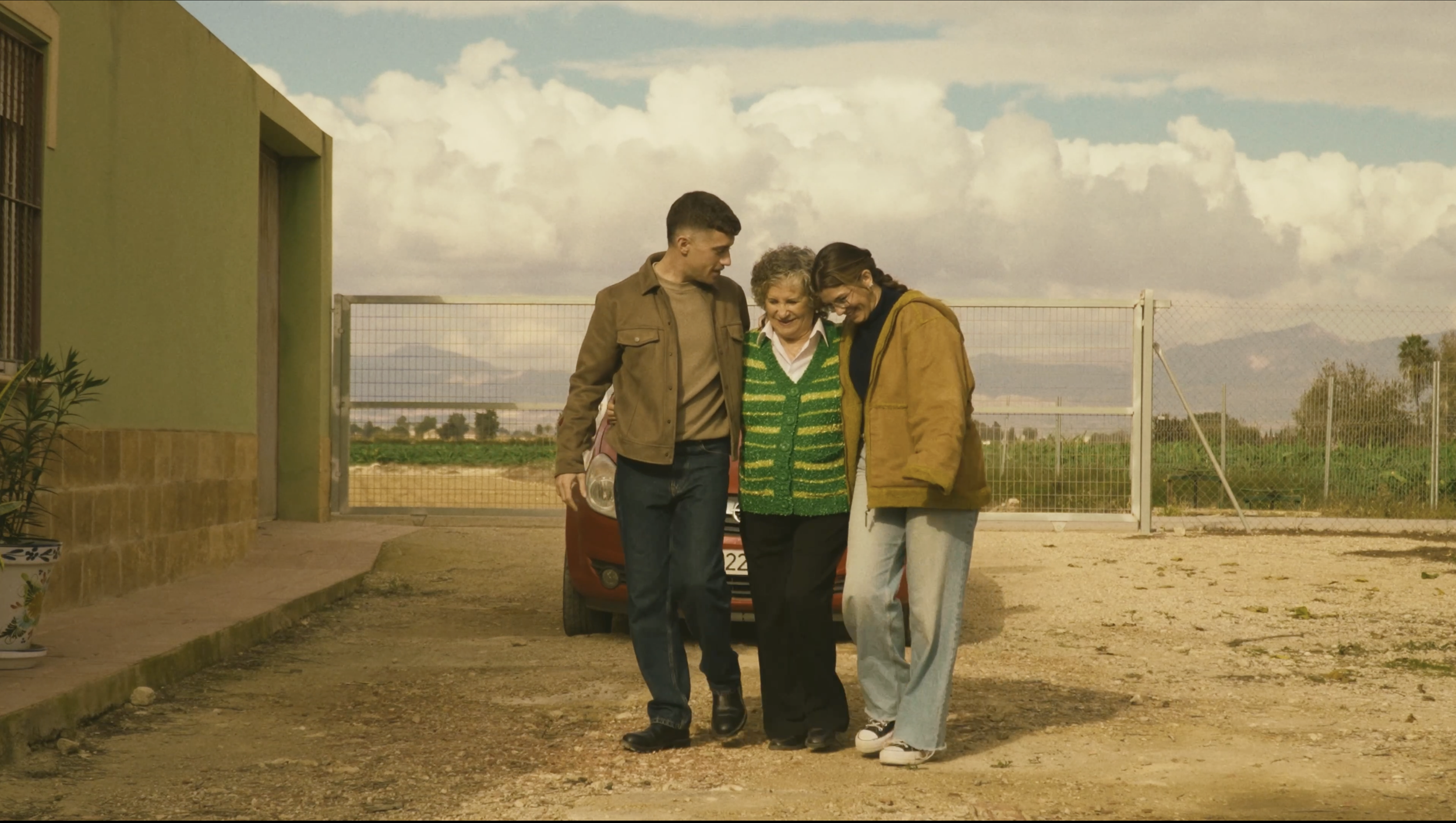 Un grupo de tres personas, dos adultos y una anciana, caminando juntos en un camino de tierra en un exterior rural, parecen felices y apoyarse mutuamente, con un coche estacionado detrás de ellos.