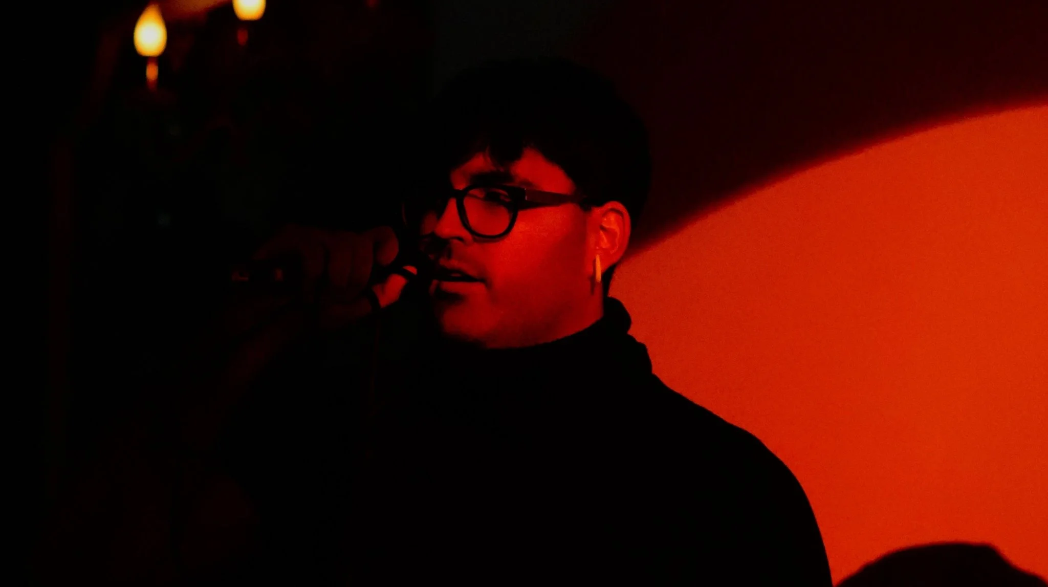 Persona con gafas y pendiente en la oreja, hablando por micrófono en un ambiente oscuro con iluminación roja.