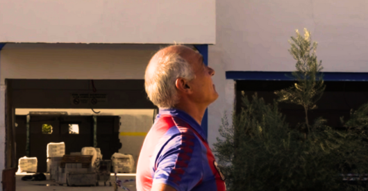 Un hombre mayor de perfil en camiseta deportiva, mirando hacia arriba, con fondo de edificio y plantas.