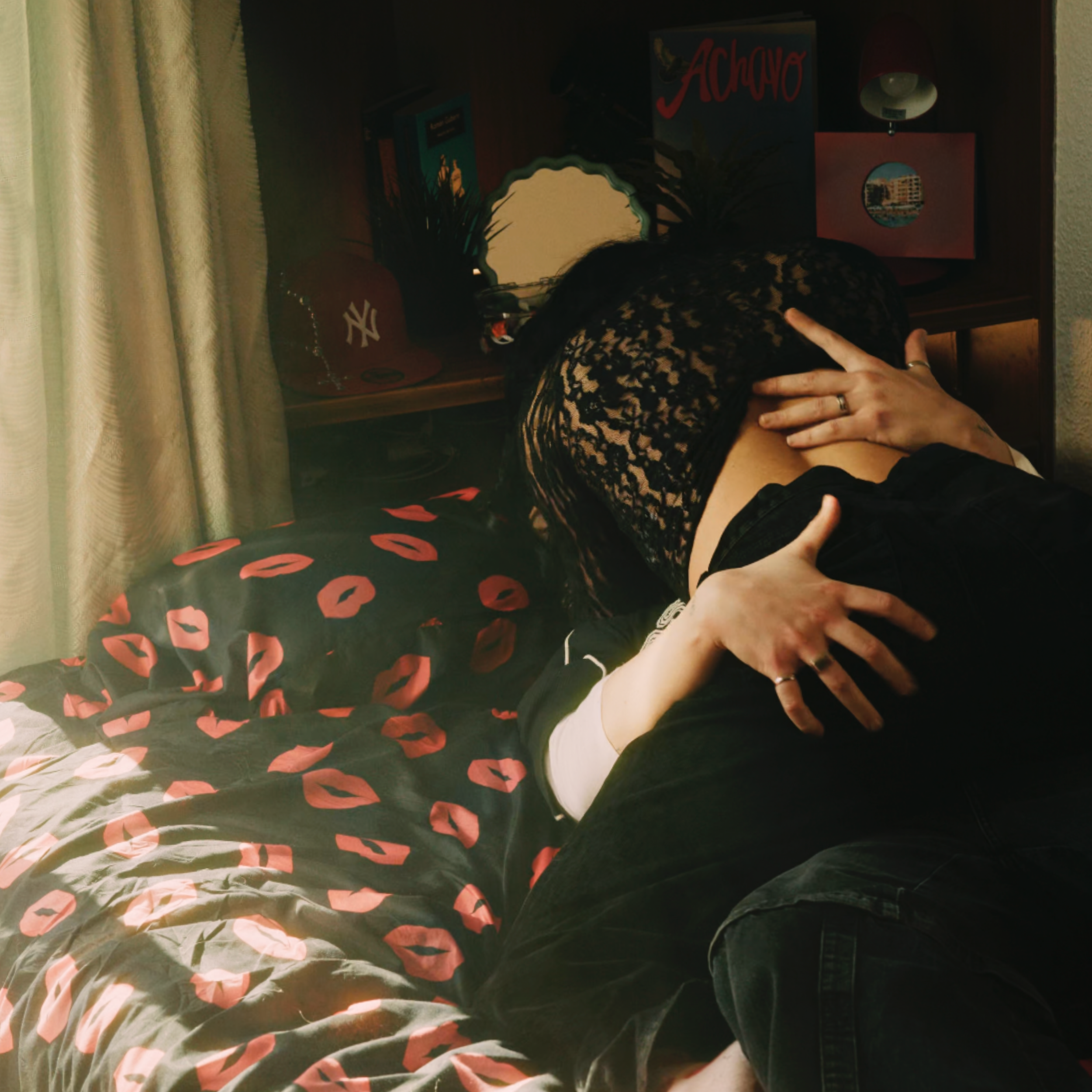 Dos personas abrazándose en la cama, con un fondo de libros y decoración en una habitación, con cortinas y una almohada con estampado de labios rojos.