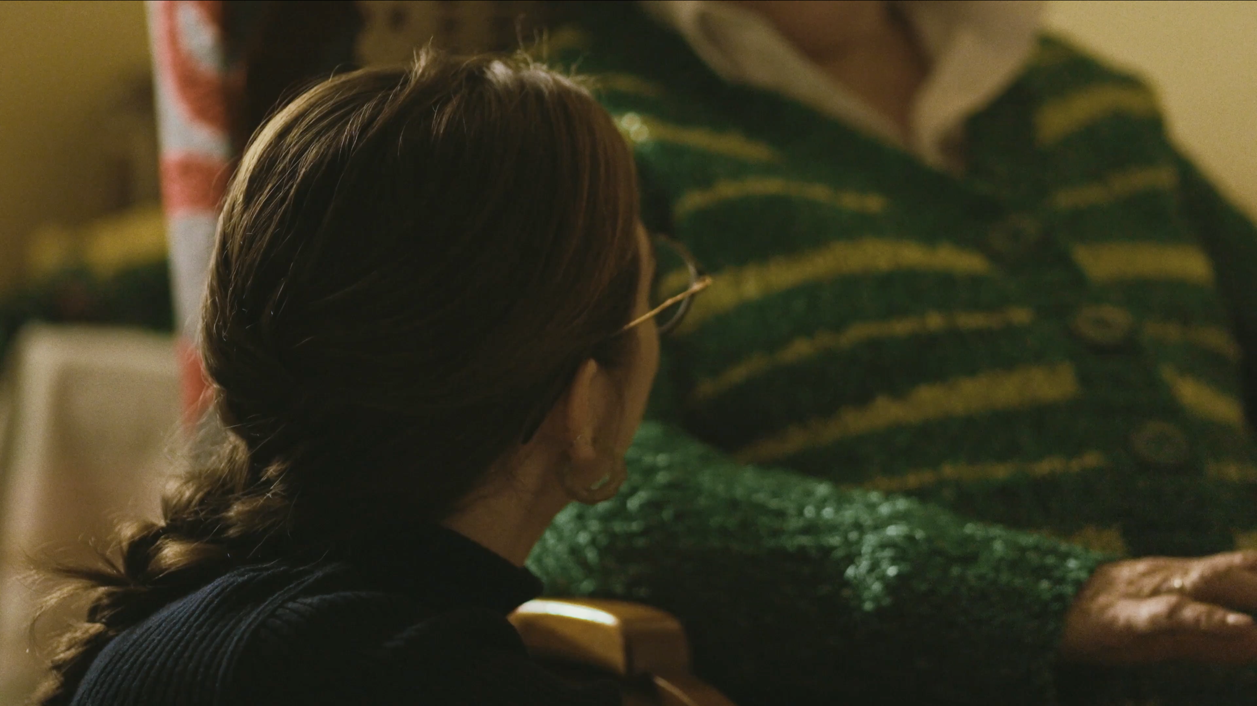 Mujer con el cabello en una trenza, usando gafas, en un ambiente con una persona mayor con ropa de rayas verdes y amarillas.