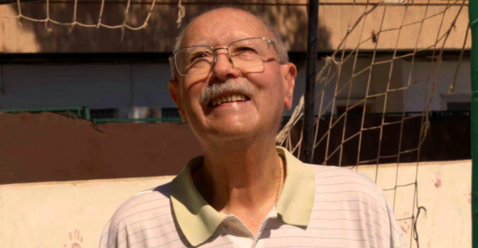Hombre mayor sonriendo en un campo de fútbol con portería de fondo.