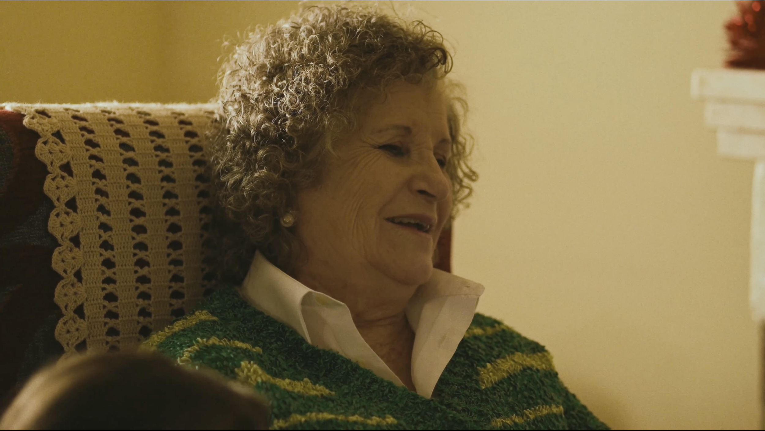 Mujer mayor con cabello rizado y canoso, sonriendo, sentada en una silla con una manta de ganchillo y ropa cómoda en un ambiente cálido.