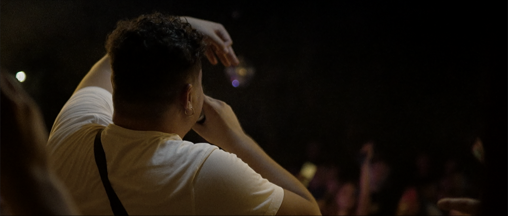 Persona con cabello rizado y cuero de manga corta, sentada en público en un lugar oscuro, funcionando como espectadora en un evento o concierto.