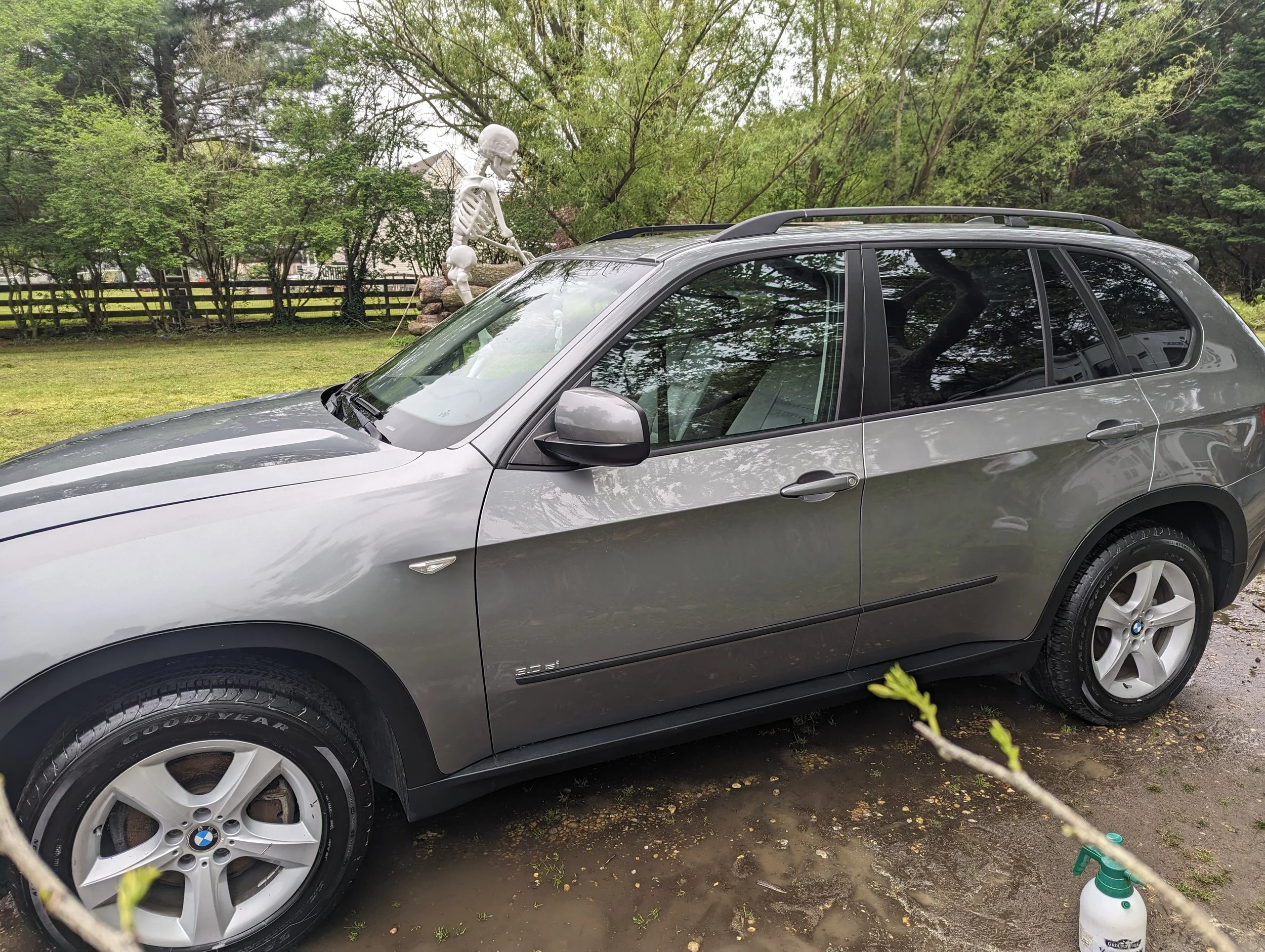 Gray BMW SUV parked on grass with trees and a white fence in the background, and a skeleton decoration on a tree in the yard.