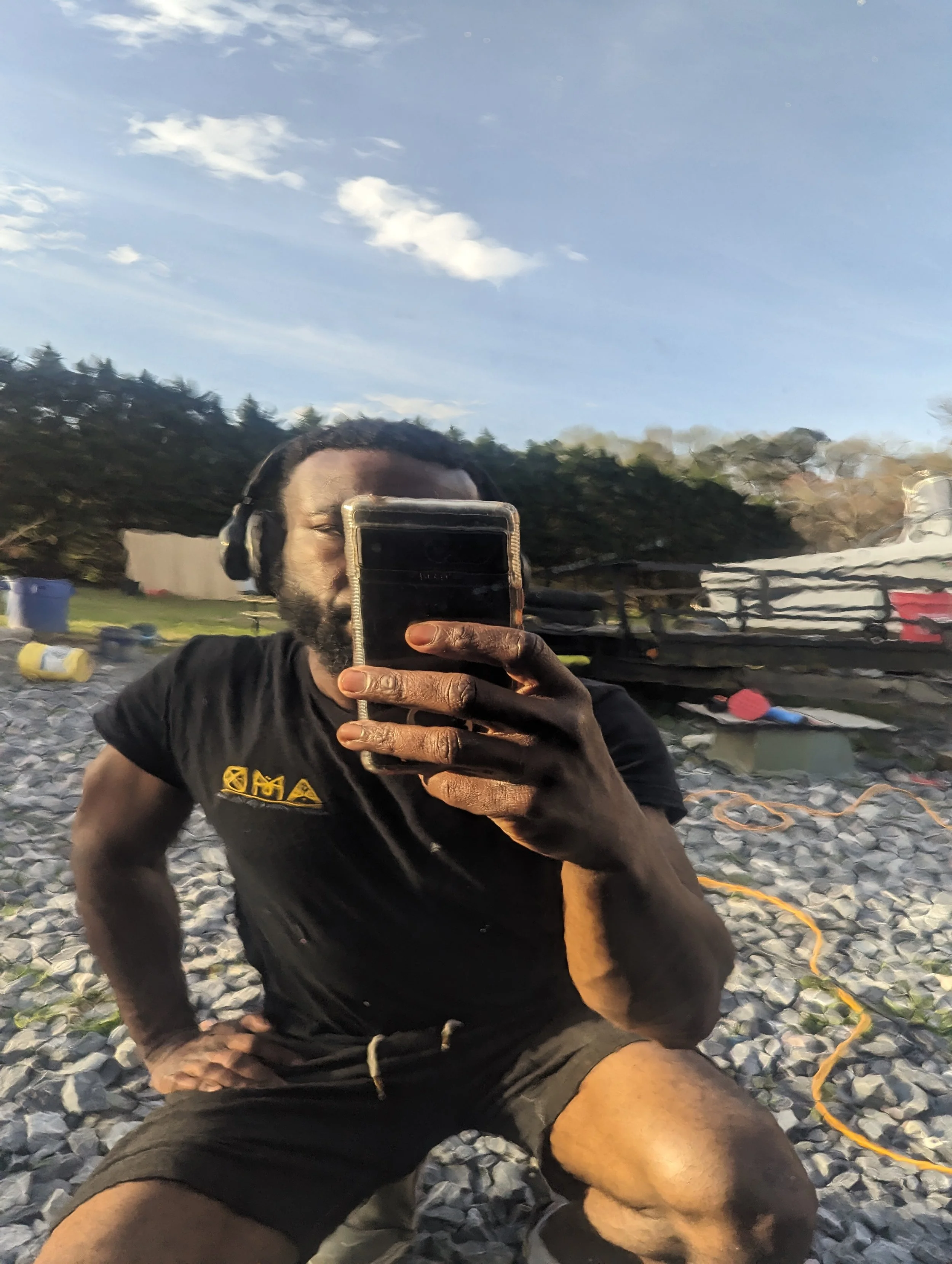 Man taking a selfie outdoors on a rocky surface with trees and equipment in the background, wearing headphones and a black T-shirt.