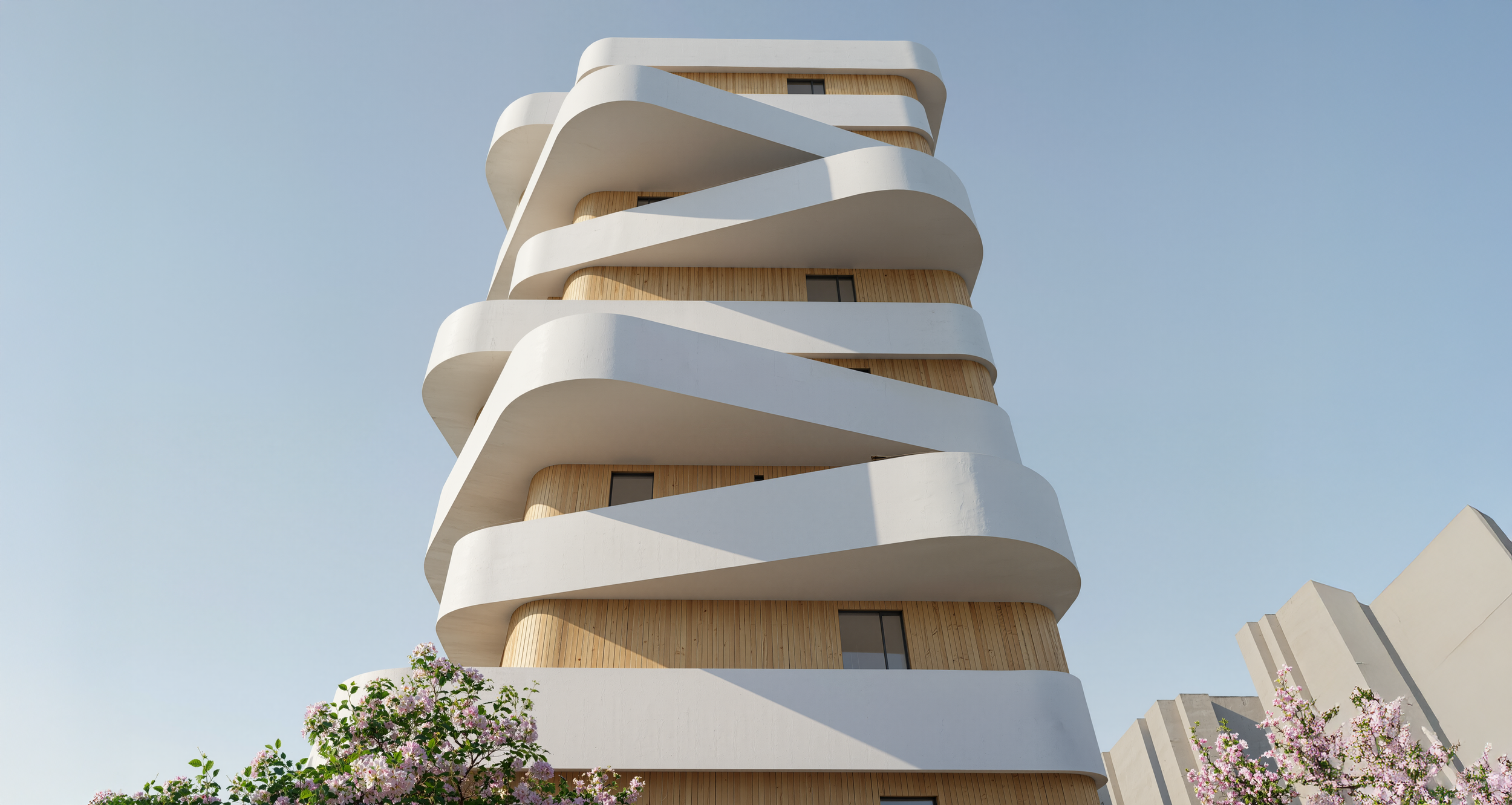 Edificio moderno de varias plantas con balcones curvos blancos y estructura de madera, rodeado de árboles con flores rosadas y un cielo despejado.