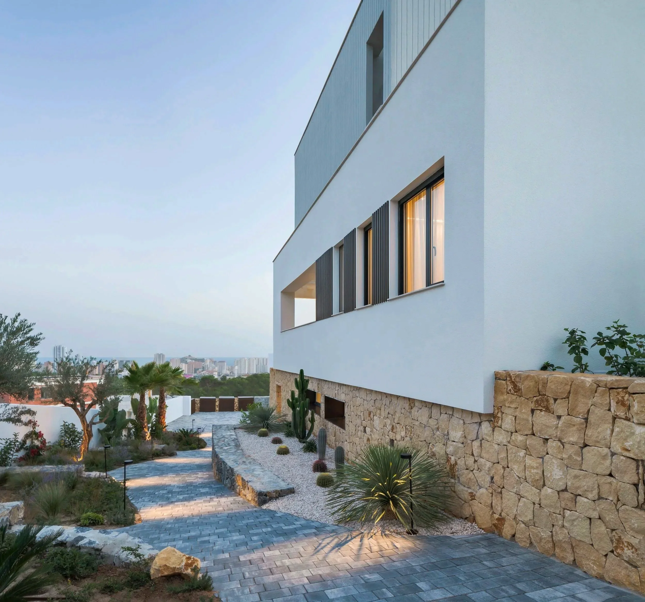 Vista de una casa moderna con paredes blancas, ventanas con persianas negras, y un jardín con plantas y palmeras en un paisaje urbano