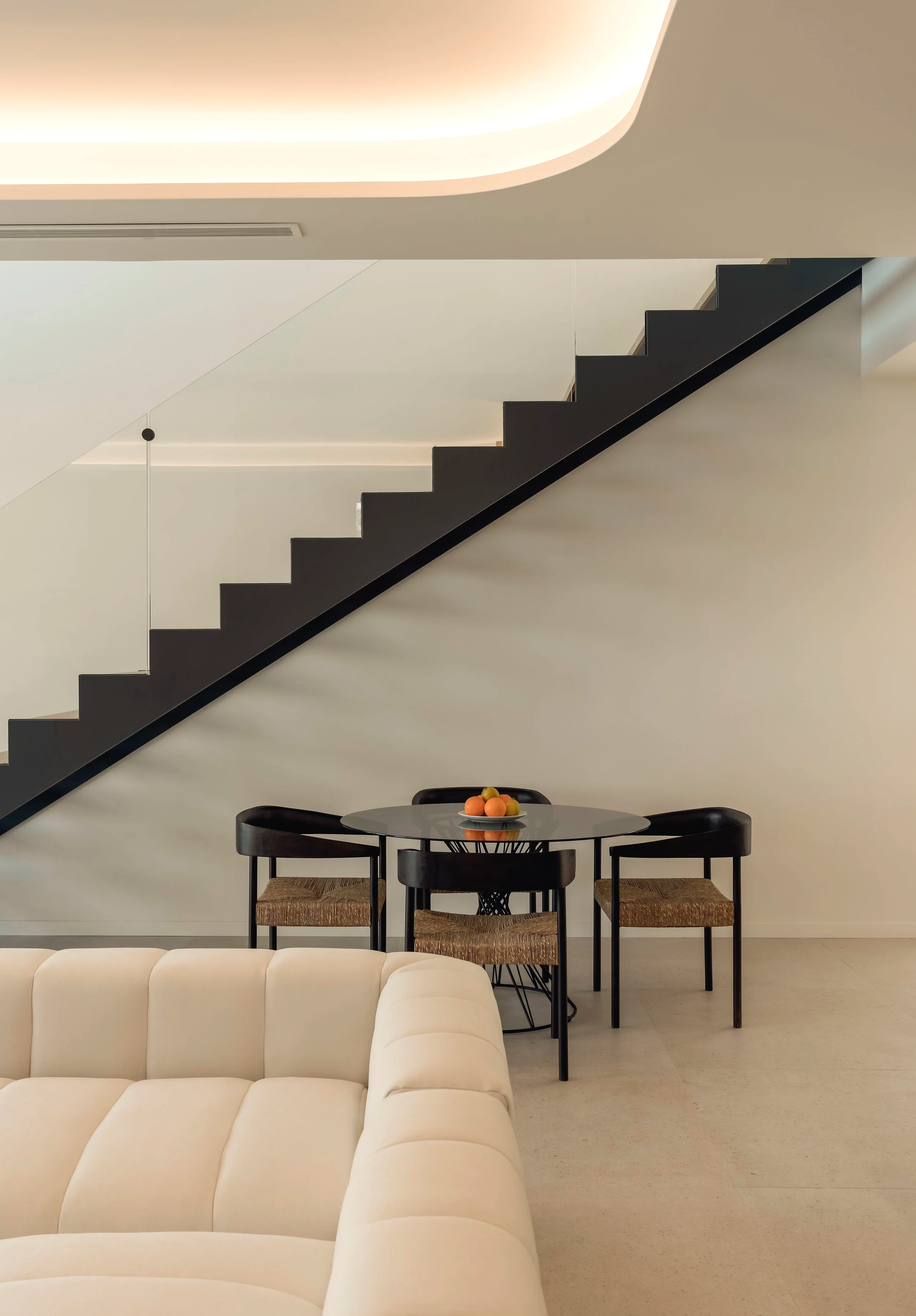 Sala moderna con una mesa redonda negra, cuatro sillas negras con asientos de tela entrelazada, y un sofá beige en primer plano, en un fondo con escalera negra y pared blanca con iluminación ambiental.