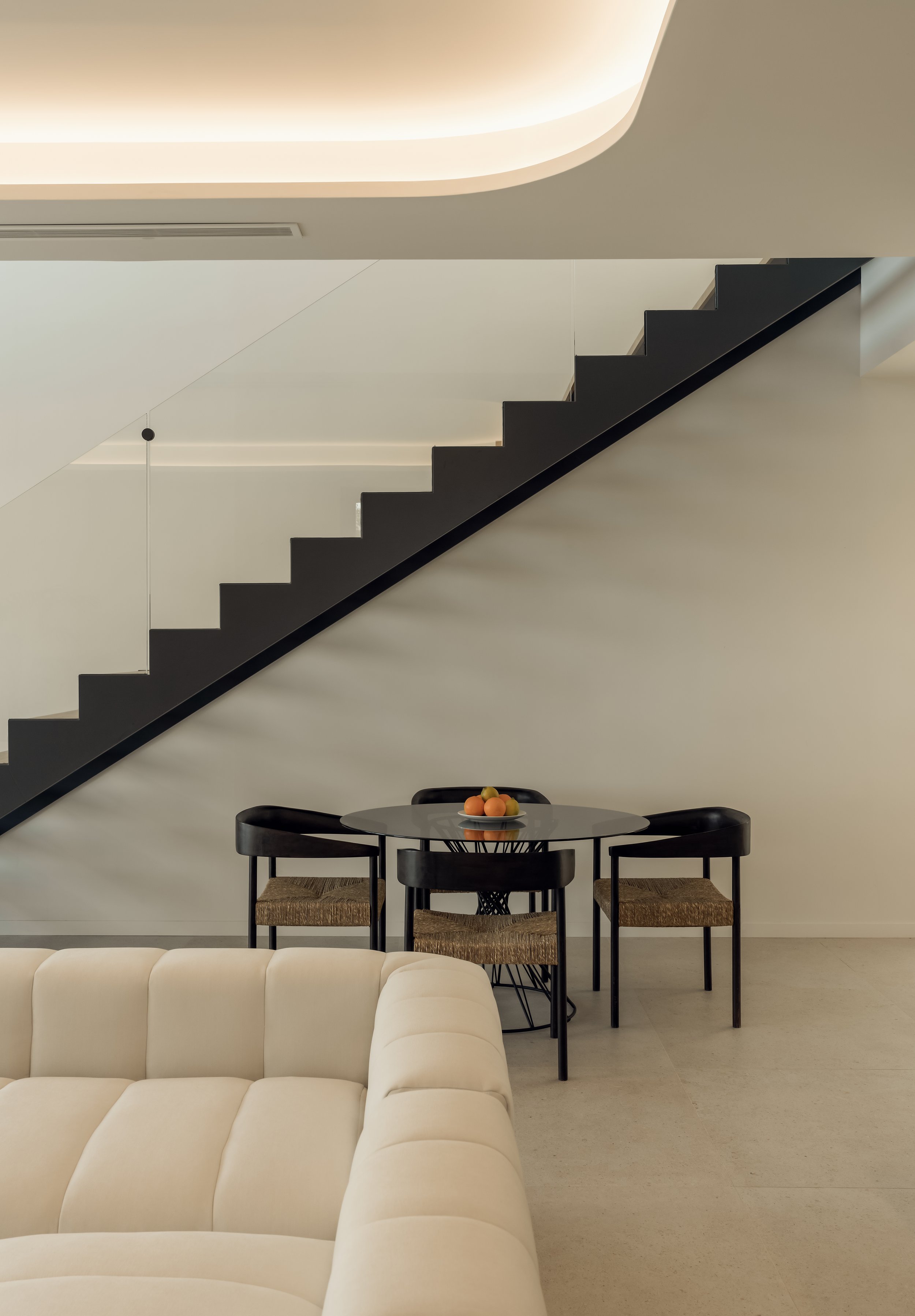 Sala moderna con una escalera negra y un sofá beige, mesa redonda con frutas y cuatro sillas negras con asientos de mimbre.