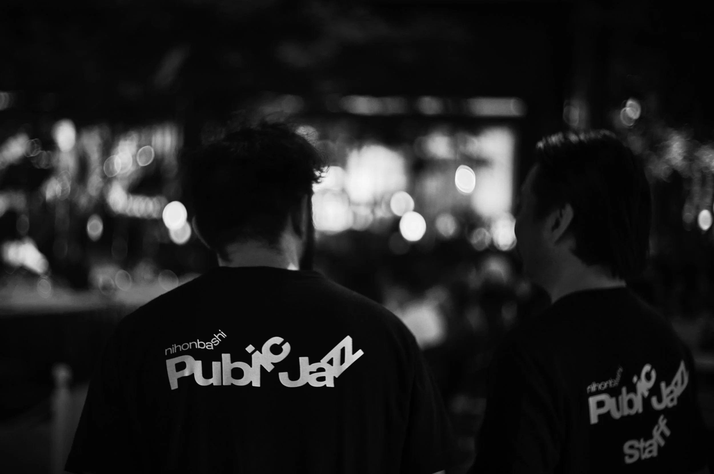 夜の都市で背中を向けた男性と女性が話している様子。男性はTシャツに「PubbJk」などと書かれている。