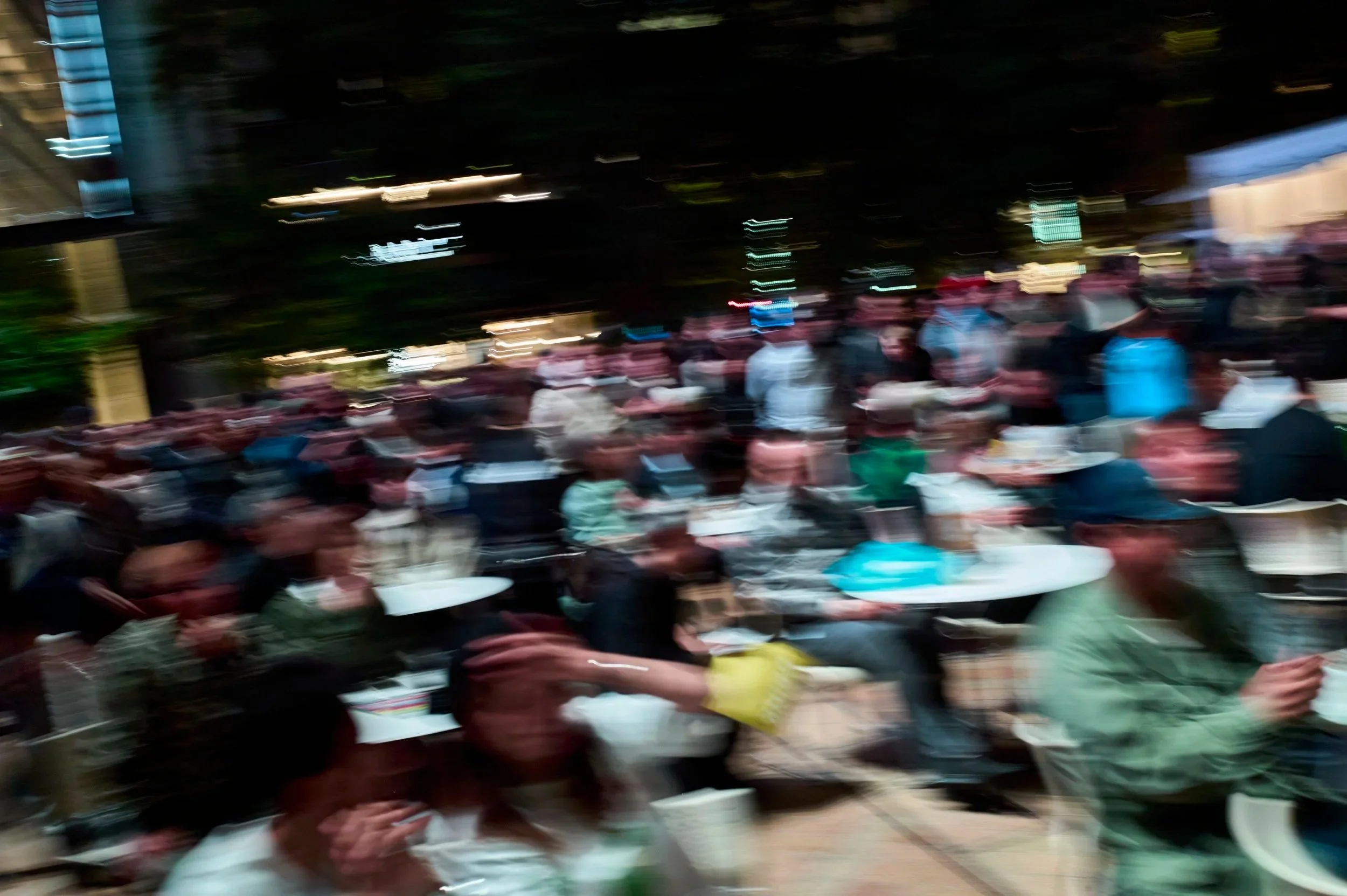 夜の屋外イベントで多くの人々が座っており、照明と建物の光がぼやけている。