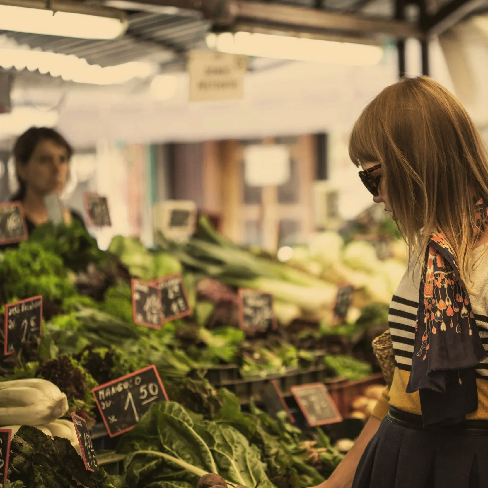 A woman wearing sunglasses shopping for vegetables at an outdoor market, with various leafy greens and produce displayed on tables.