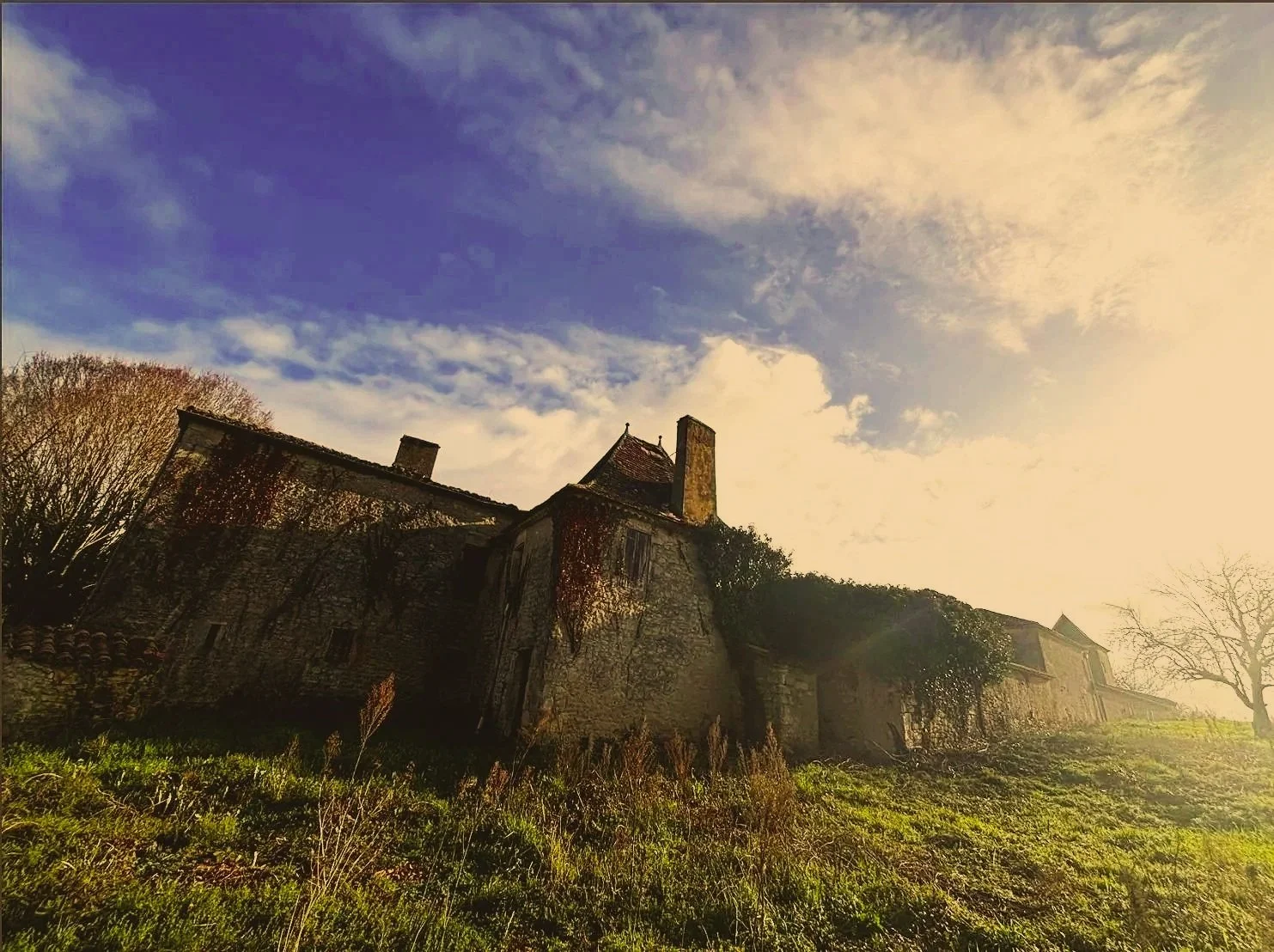 An old stone house with ivy growing on the walls, surrounded by grass and trees, under a partially cloudy sky during sunset or sunrise.