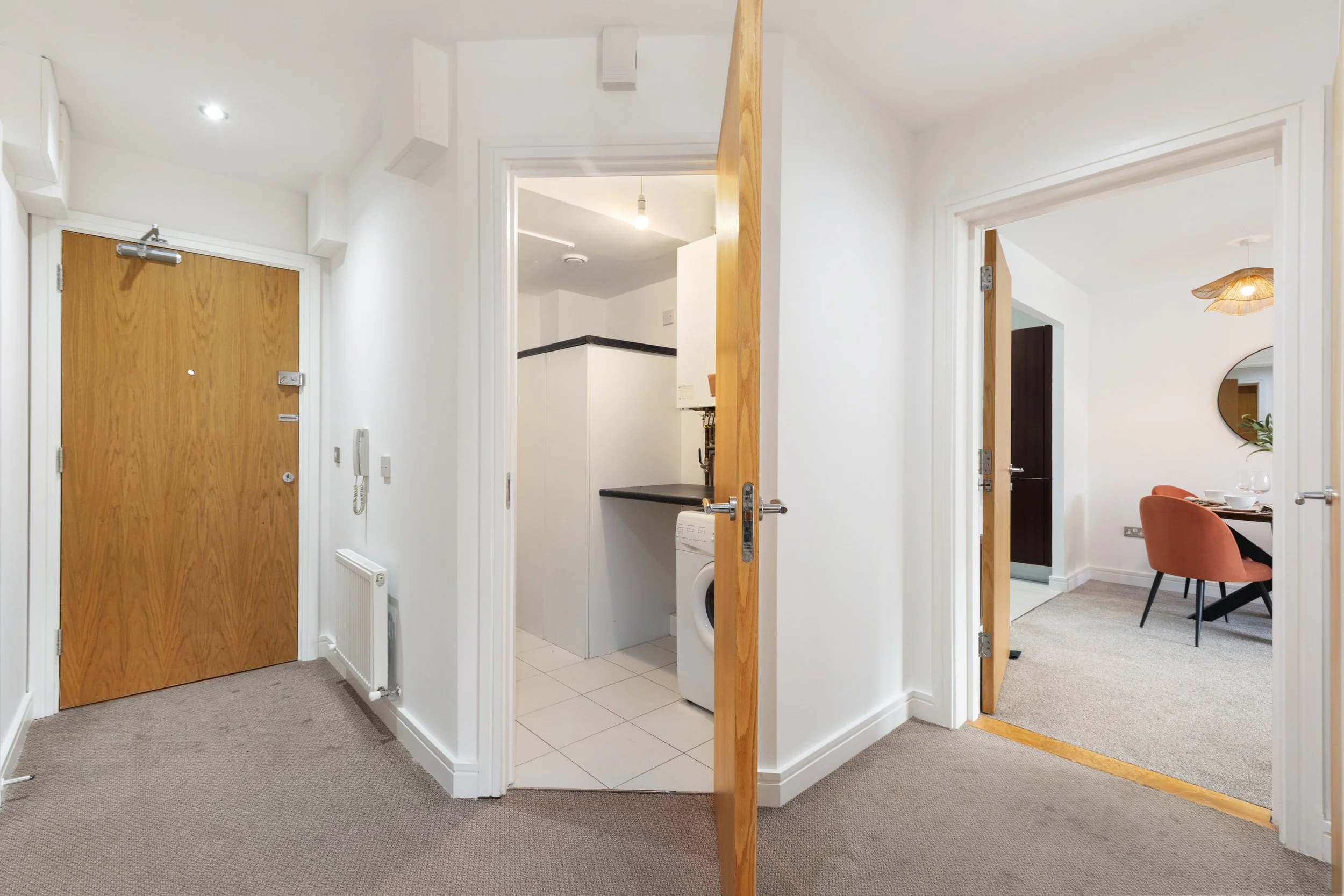 Interior view of an apartment hallway with a wooden front door on the left, a laundry area with a washing machine in the center, and a dining room with a round mirror and orange chairs on the right.