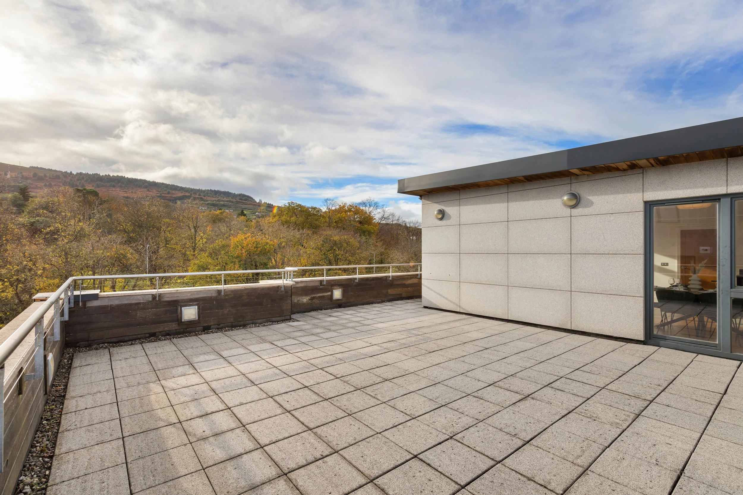 Empty rooftop terrace with tiled floor, modern building wall, railing, scenic fall landscape with trees and mountains in the background, partly cloudy sky.