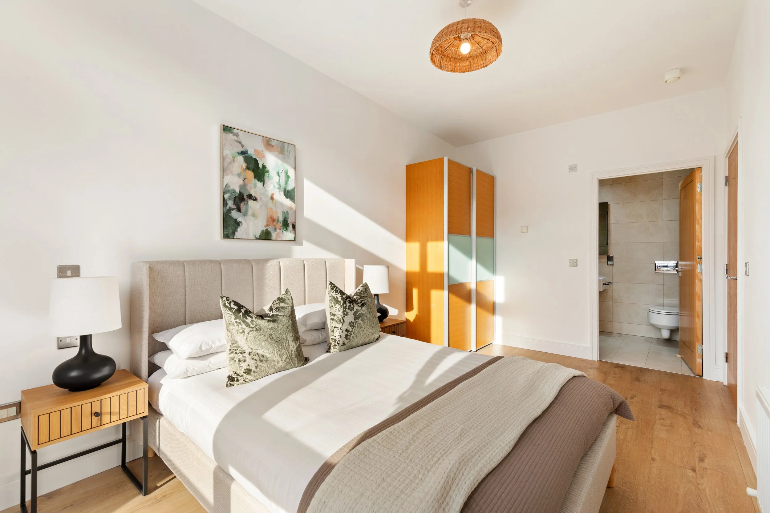 Hotel bedroom with a bed, two nightstands with lamps, a colorful abstract painting, a wooden wardrobe, and an open door leading to a bathroom with beige tiles.