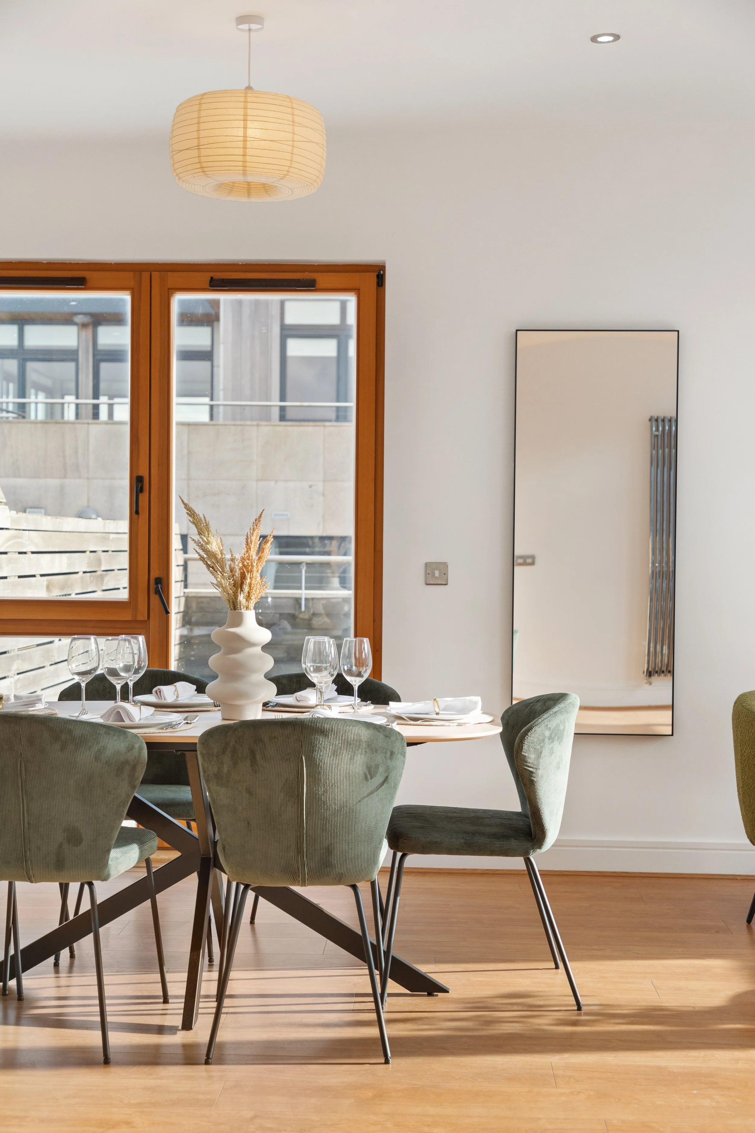 Dining room with a table set for four, green upholstered chairs, a white vase with dried pampas grass, wooden flooring, and large glass doors leading outside.