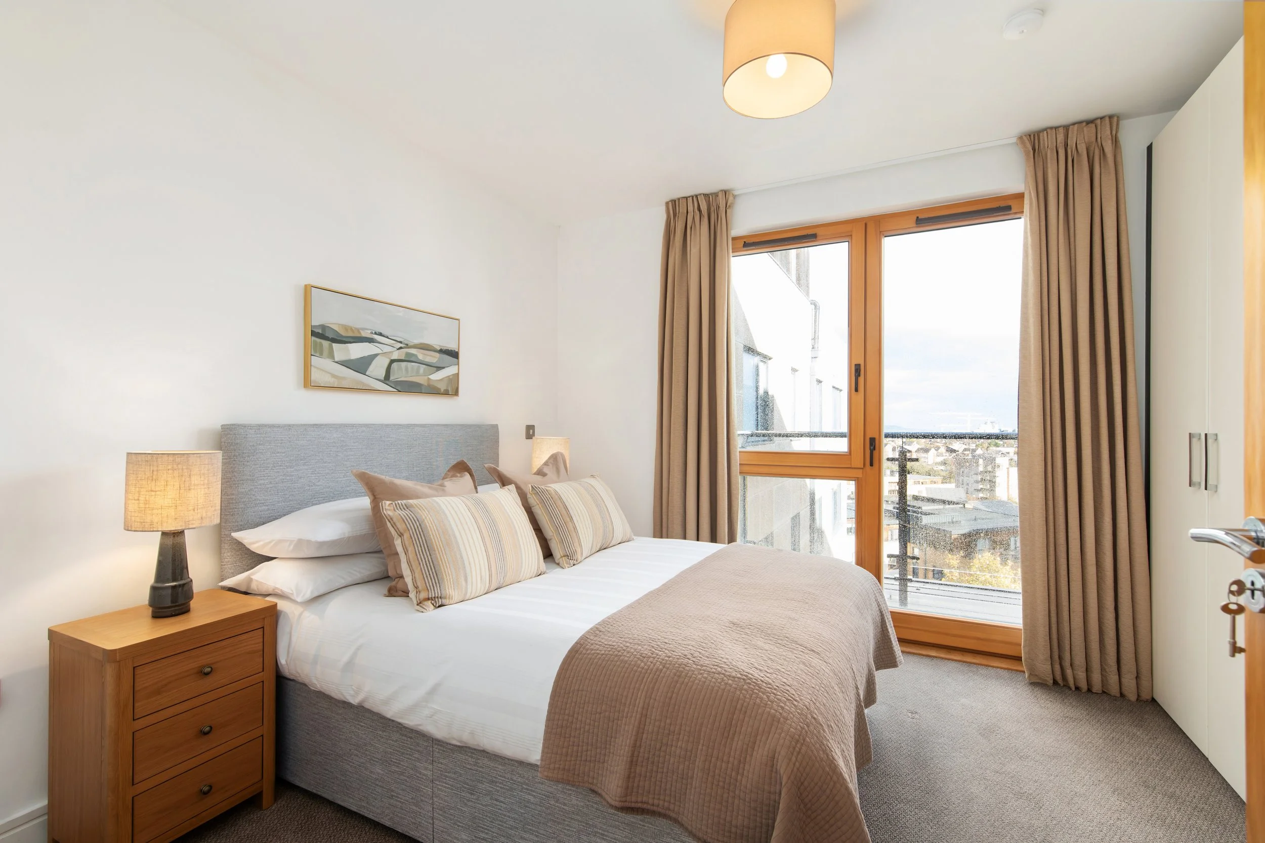 A bedroom with a gray upholstered bed, beige bedding, and striped pillows. There is a wooden nightstand with a lamp on it, and a sliding glass door with beige curtains leading to a balcony. A painting hangs above the bed.