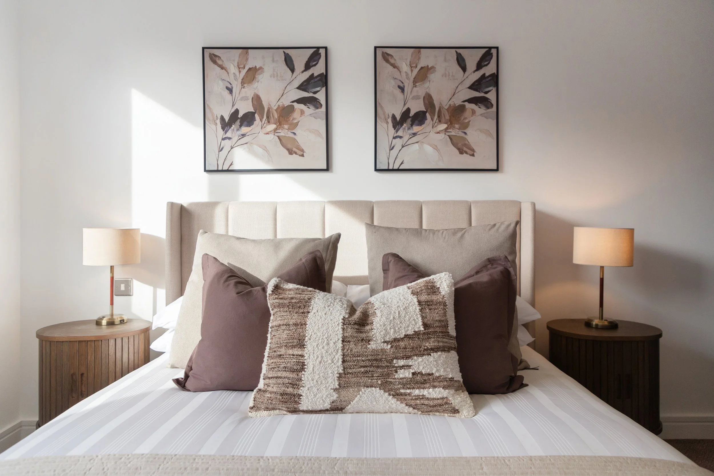 A bedroom with a beige upholstered headboard, decorated with four pillows in shades of beige and brown, with two framed botanical paintings above, and two matching bedside tables with lamps.