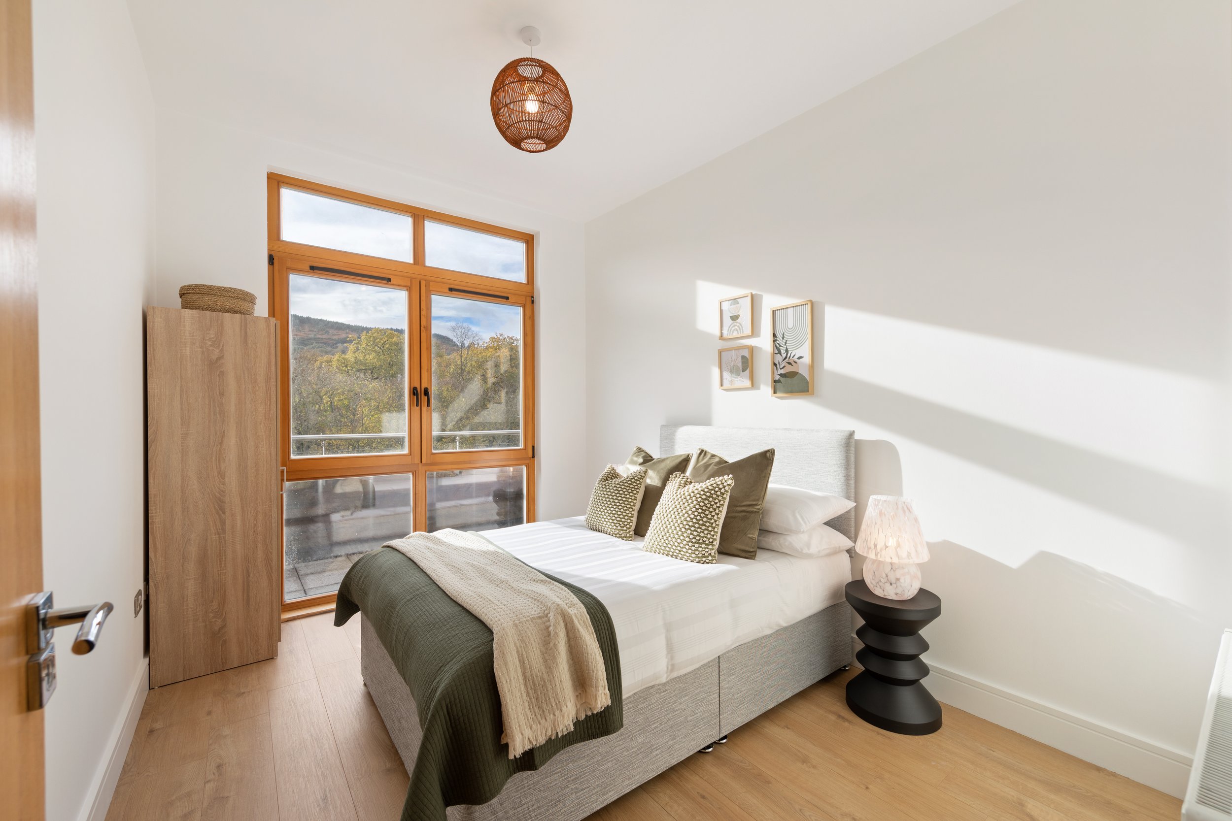 Bedroom with large window showing outdoor view, bed with neutral bedding and patterned pillows, wooden wardrobe, black side table with lamp, white walls, and minimalistic wall art.