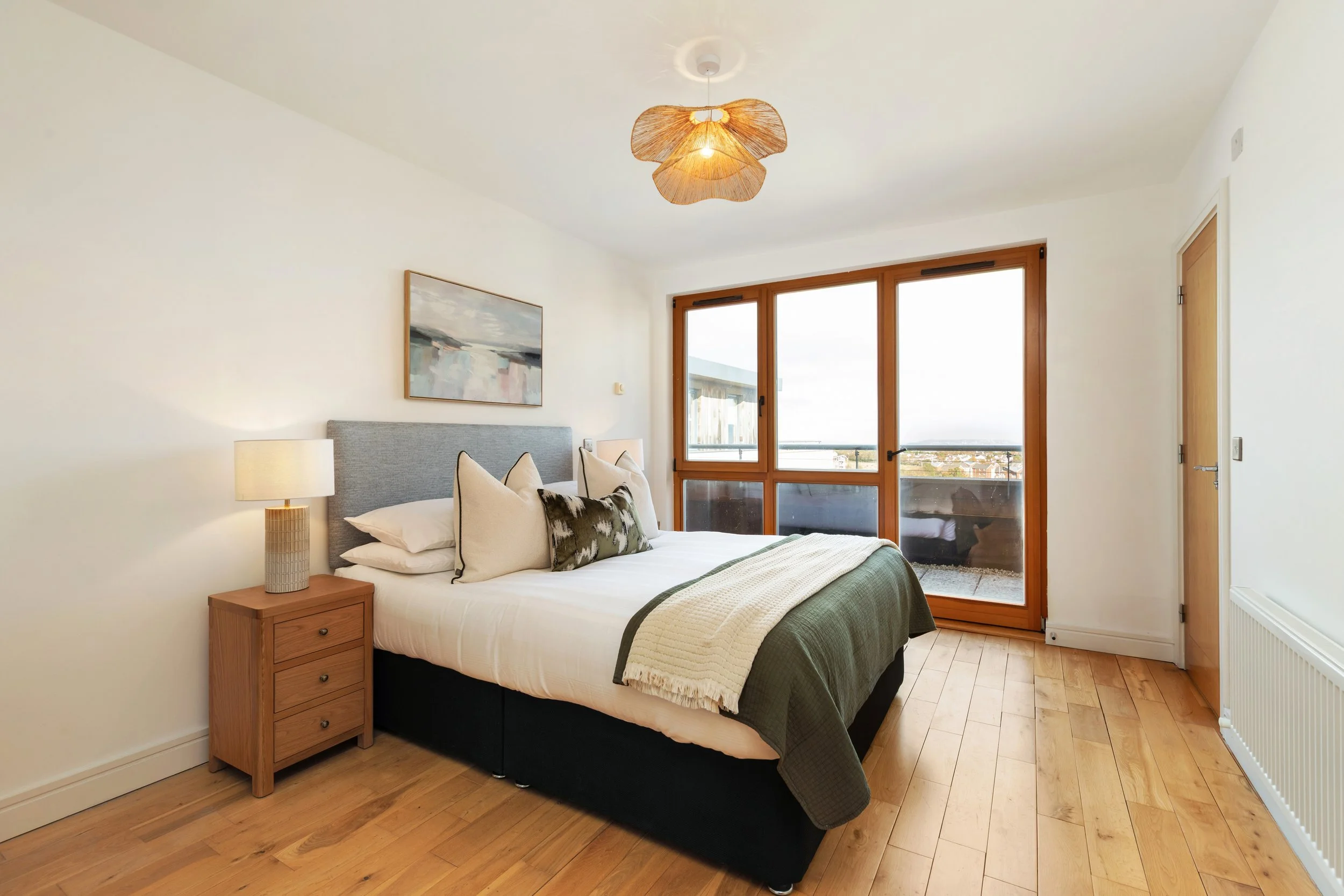 A bedroom with a bed, pillows, a nightstand with a lamp, a painting on the wall, and a large window showing a view outside. The room has wooden floors and a ceiling light fixture.