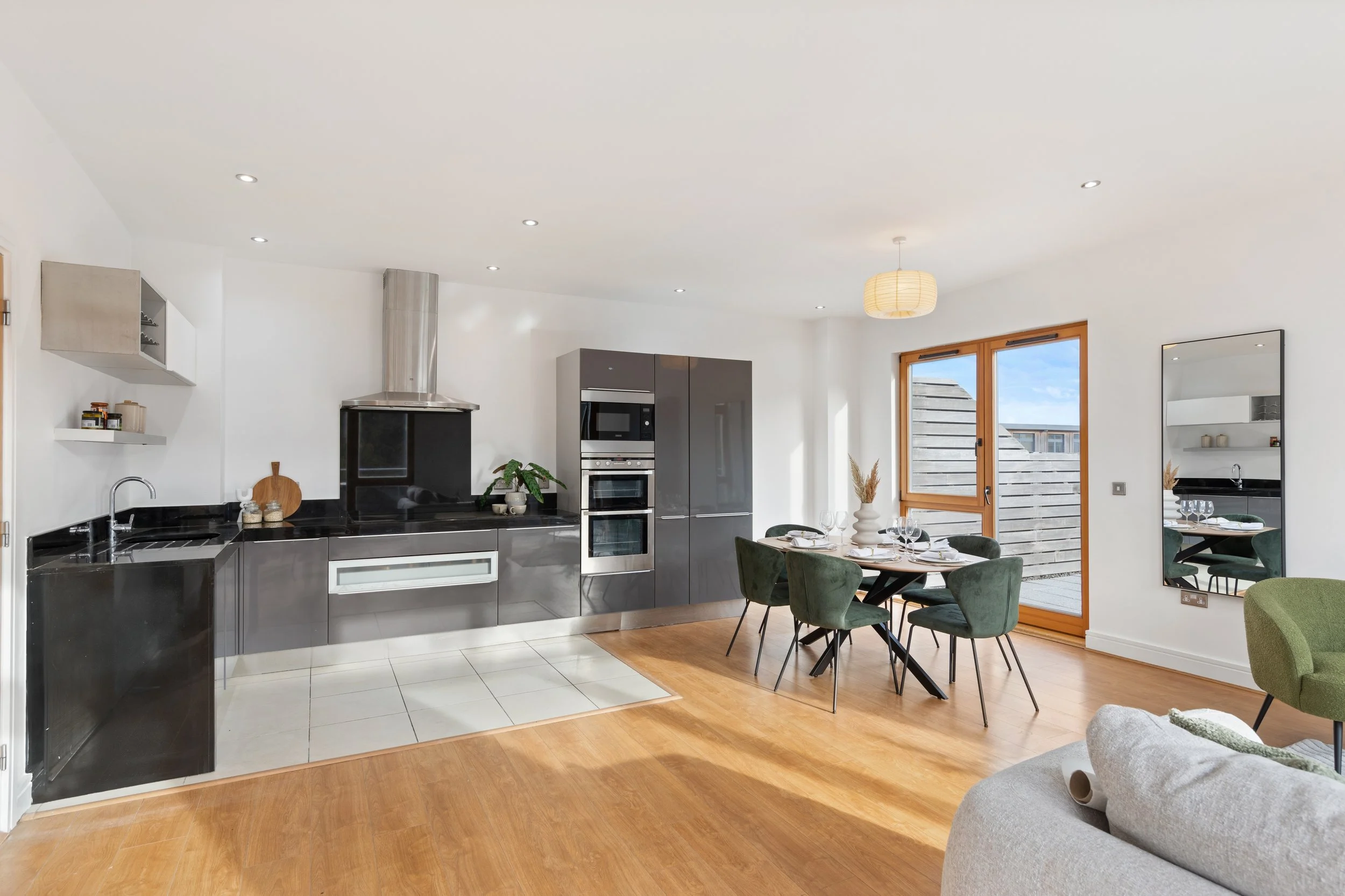 Open-plan kitchen and dining area with black and gray cabinetry, white walls, wooden flooring, and a large window letting in natural light. A round dining table with six green chairs is set for a meal. A mirror on the wall reflects part of the space.