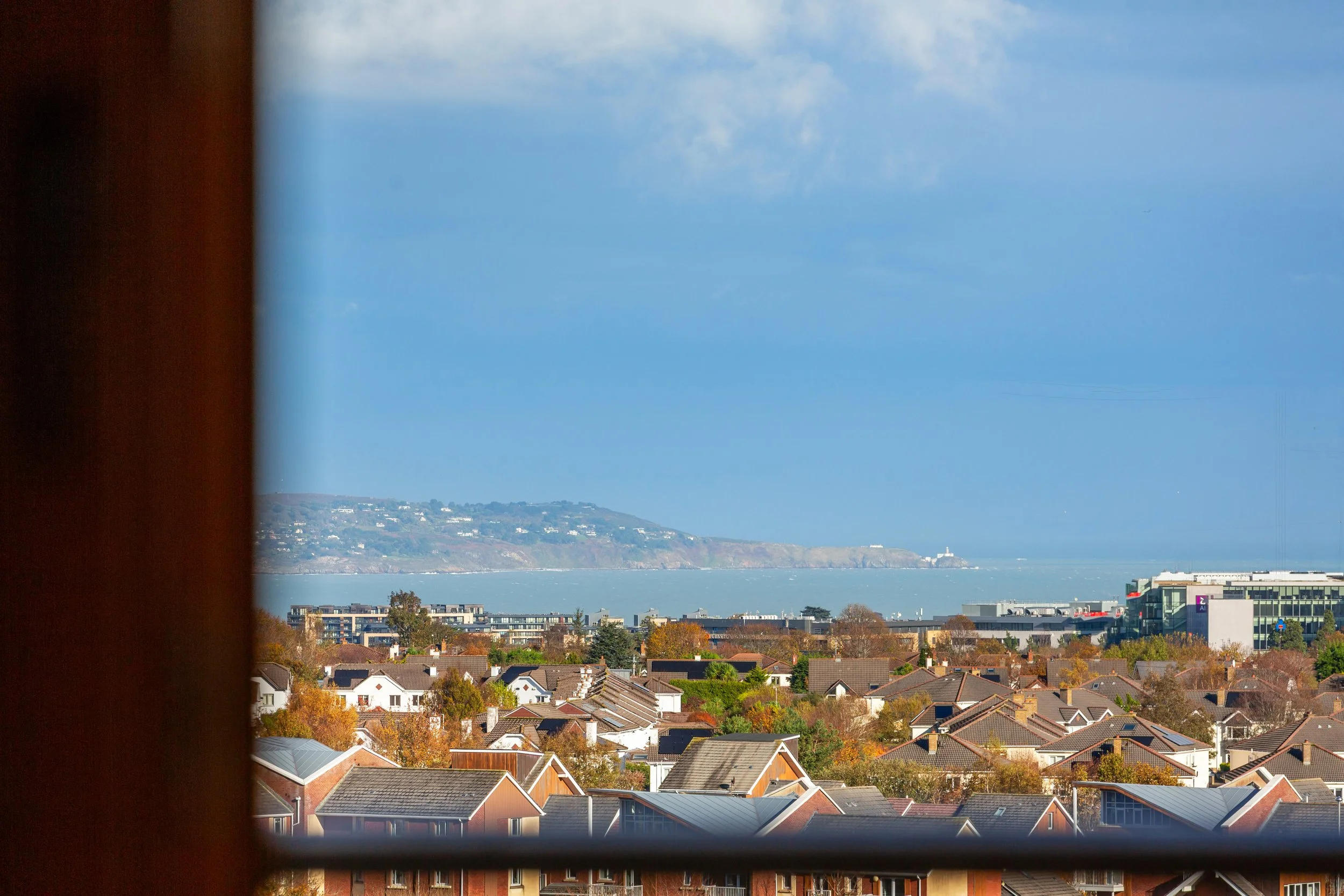 View of a cityscape with houses, trees, and buildings, overlooking a large body of water with a landmass in the distance, seen through a window with a dark curtain on the left.