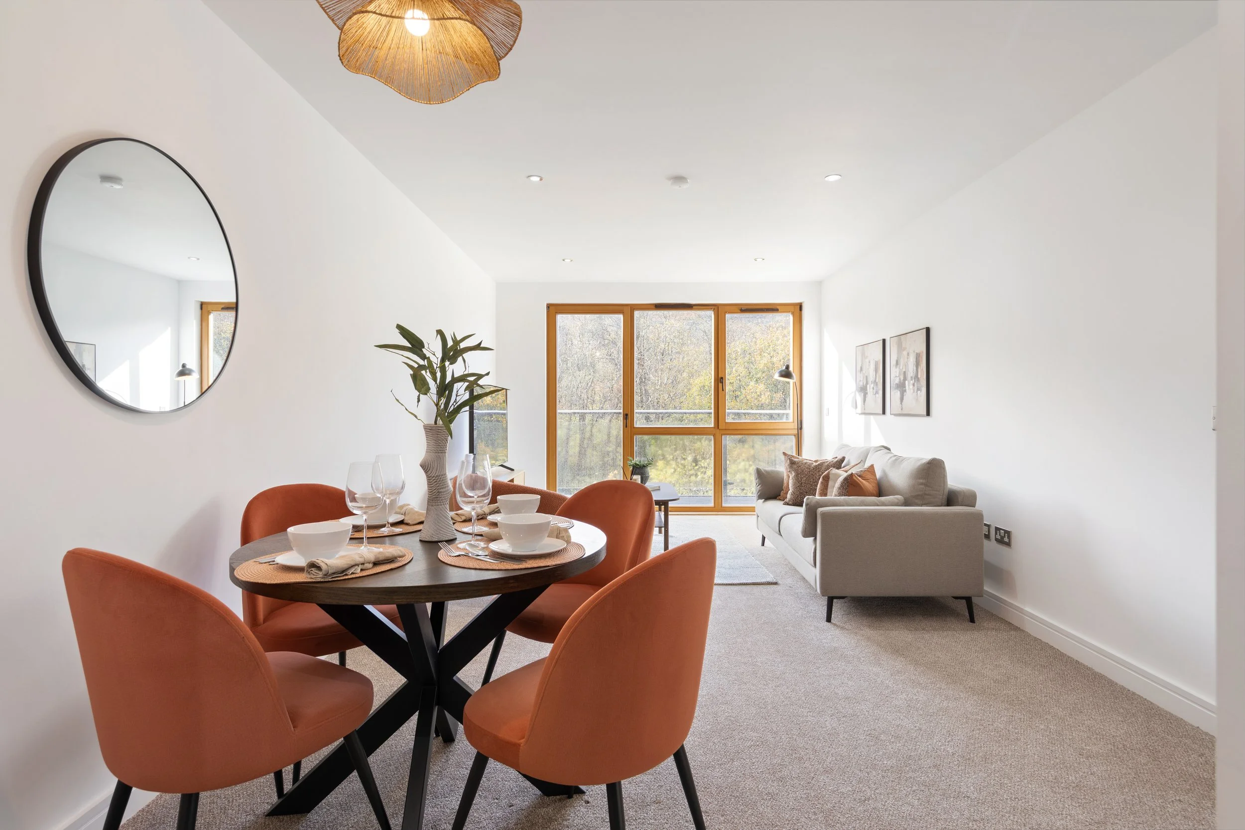 Modern living room and dining area with a round wooden table, four orange velvet chairs, a white sofa, large windows, a mirror on the wall, and decorative lamps and artwork.