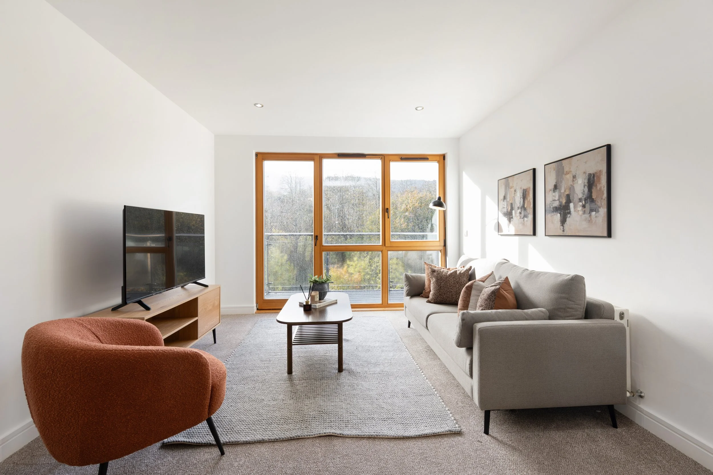 A modern living room with a light gray sofa, a rust-colored accent chair, a wooden media console with a flat-screen TV, a rectangular wooden coffee table, beige carpeting, white walls with abstract art, and large windows with wooden frames showing tr