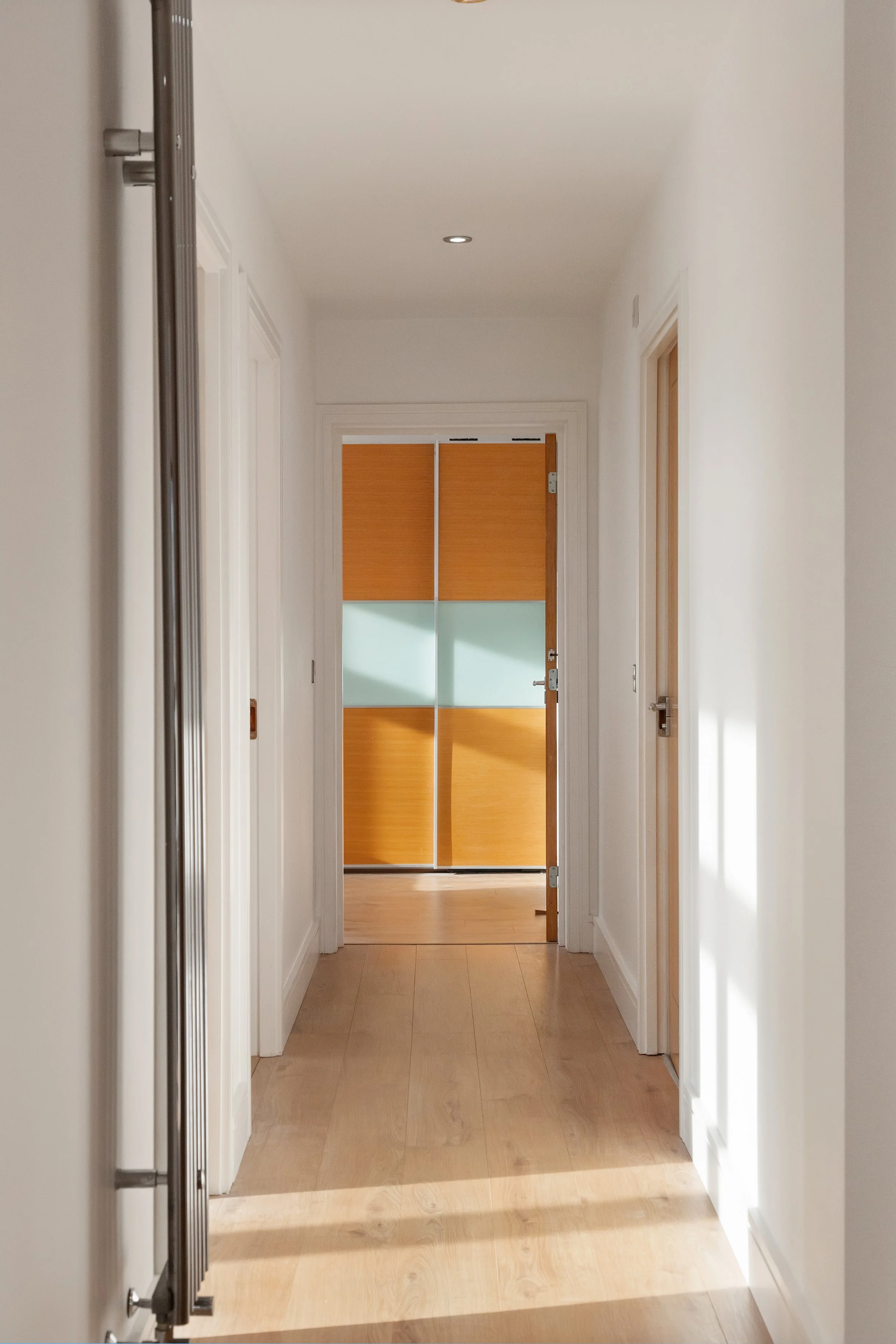 Bright hallway with light hardwood floors, white walls, and a wooden door at the end with frosted glass panels.
