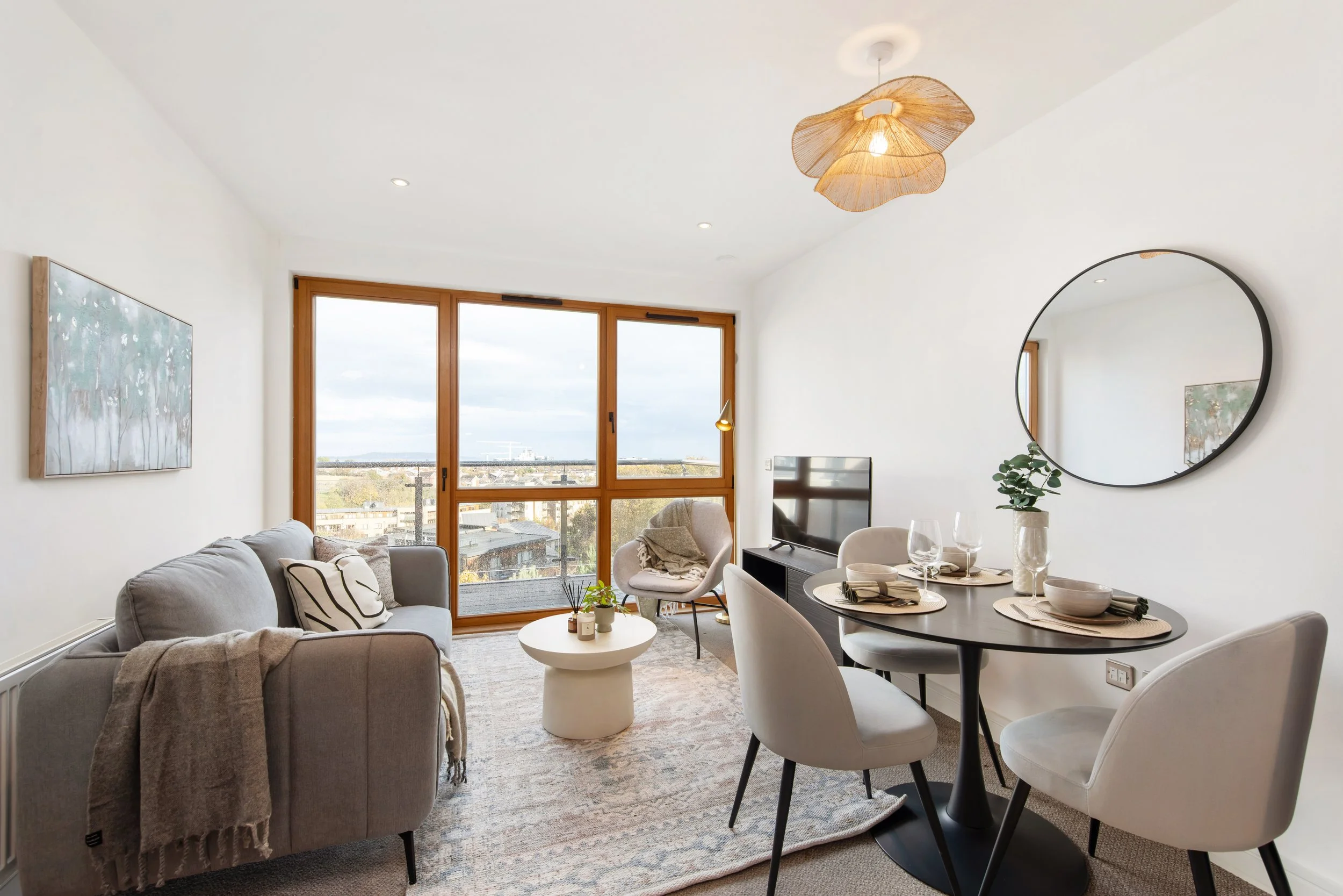Modern living room with beige sofa, armchair, round dining table set with white chairs, large window with wooden frames, wall mirror, TV, and decorative items.