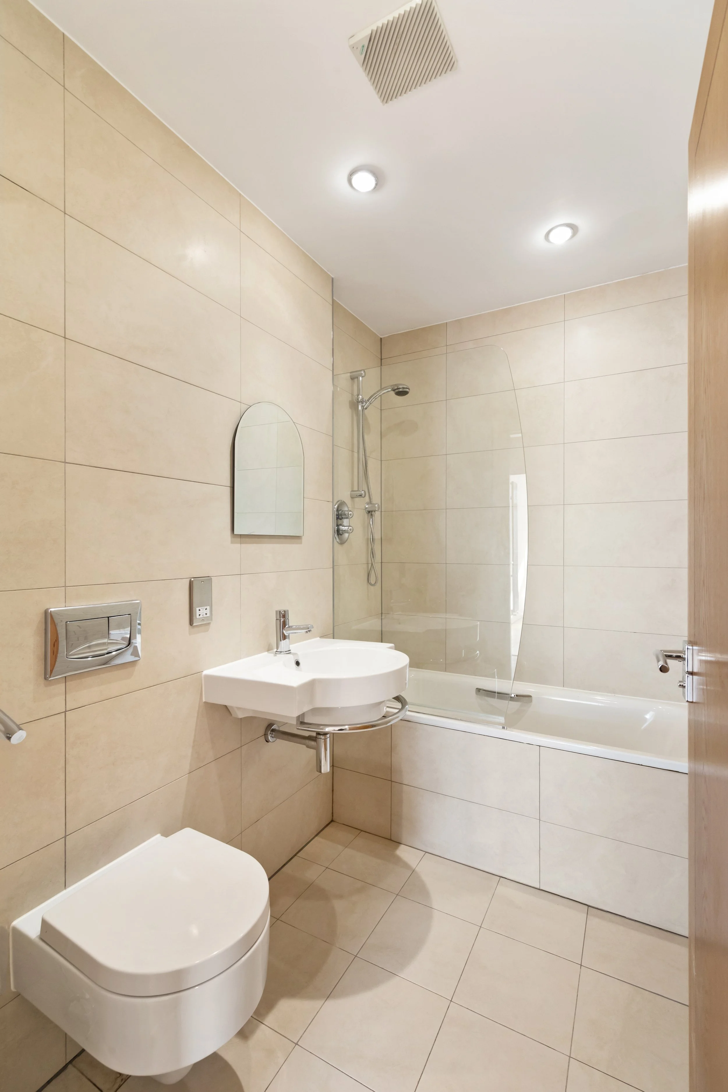 A clean, modern bathroom with beige tiled walls and floor, a white wall-mounted toilet, a small white corner sink, a mirror, a shower area with a glass screen, and ceiling-mounted lights.