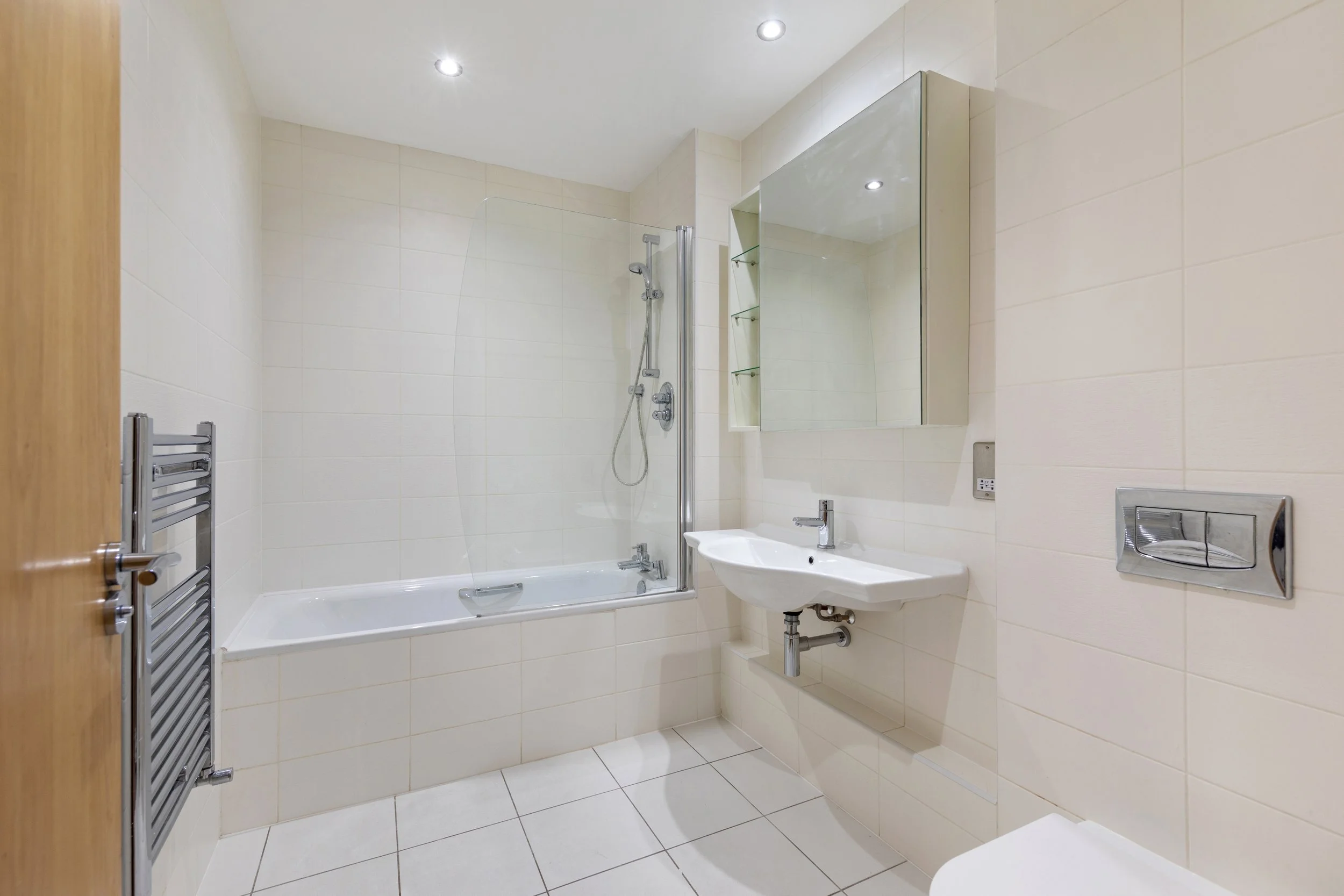 Modern bathroom with a bathtub, showerhead, white tile walls, wall-mounted sink, mirrored cabinet, and toilet, illuminated by recessed ceiling lights.