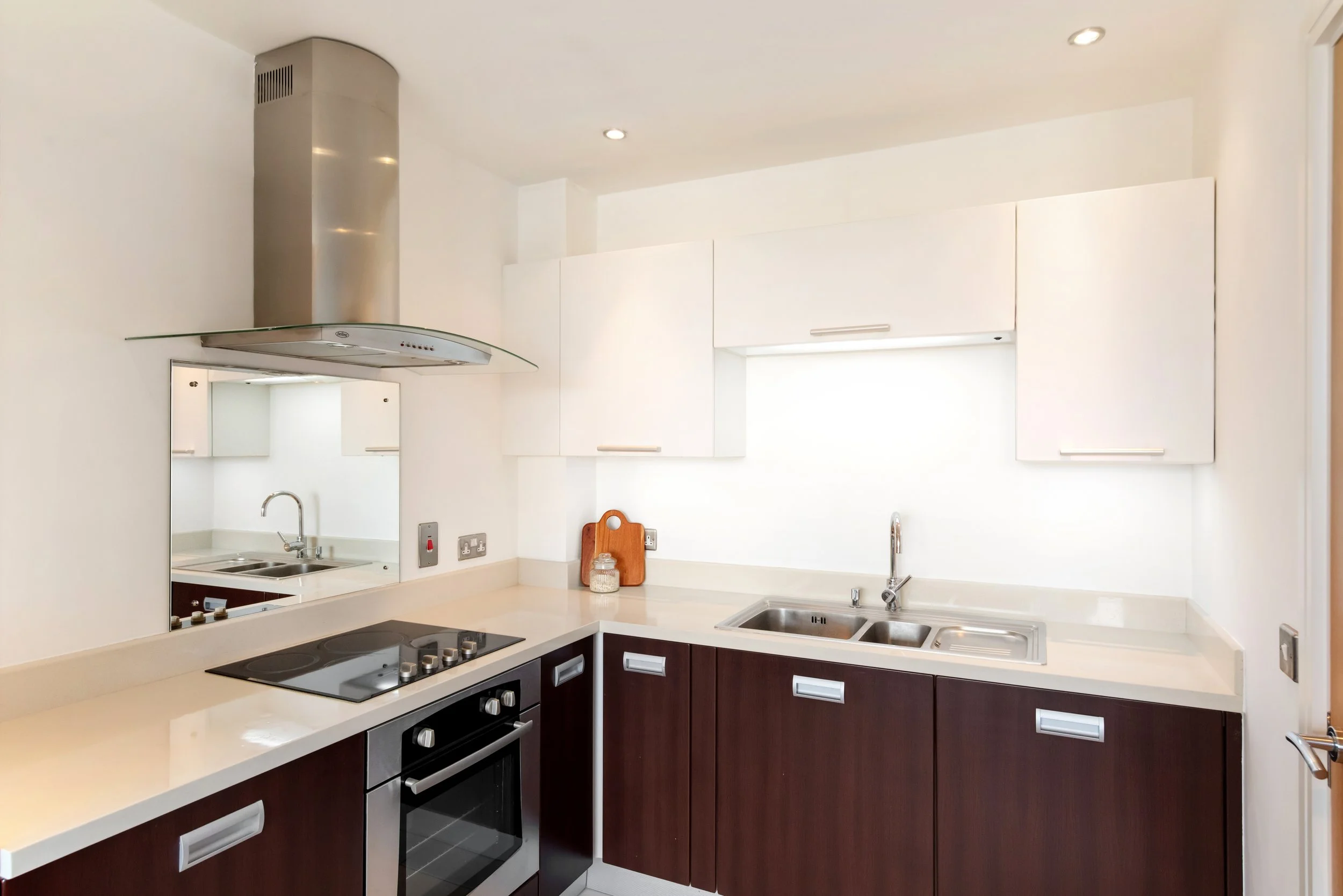 Modern kitchen with white upper cabinets, dark lower cabinets, stainless steel oven and stovetop, range hood, double sink, and a small mirror on the wall reflecting the sink and cabinets.