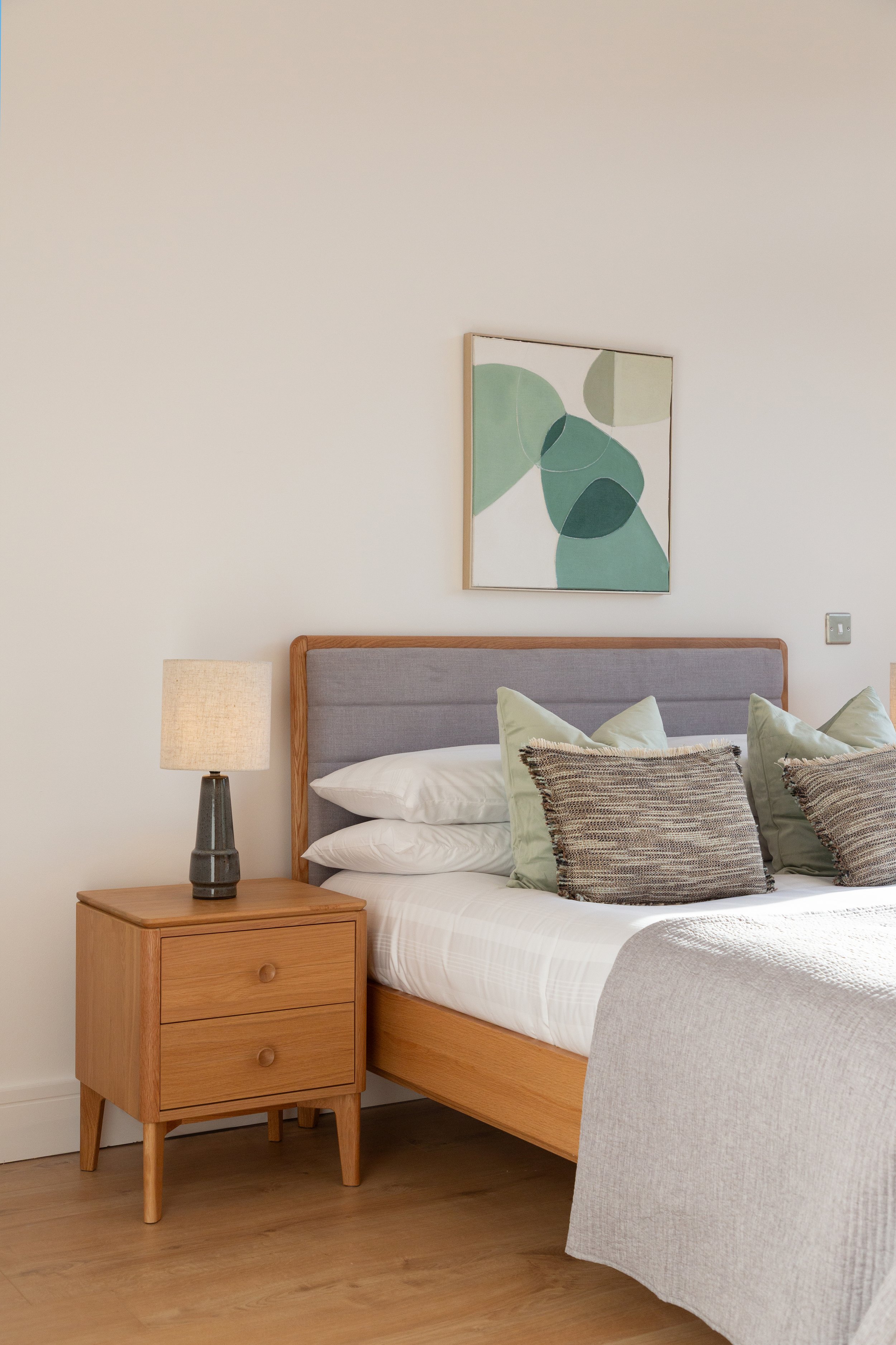 A bedroom with a wooden nightstand, a gray upholstered headboard, a table lamp, a bed with white sheets and multiple pillows, and a green abstract art print on the wall.