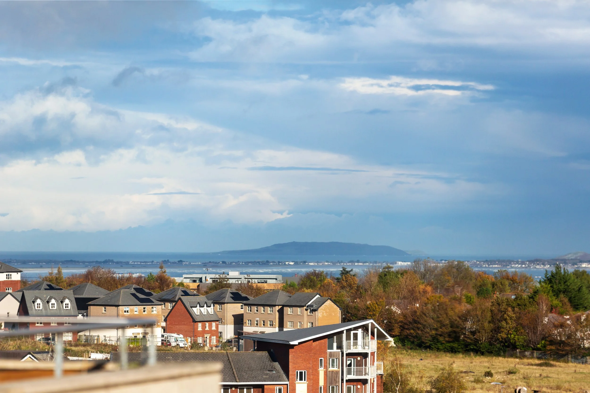 A view of a suburban neighborhood with multiple houses, trees, and a body of water in the distance under a mostly cloudy sky.