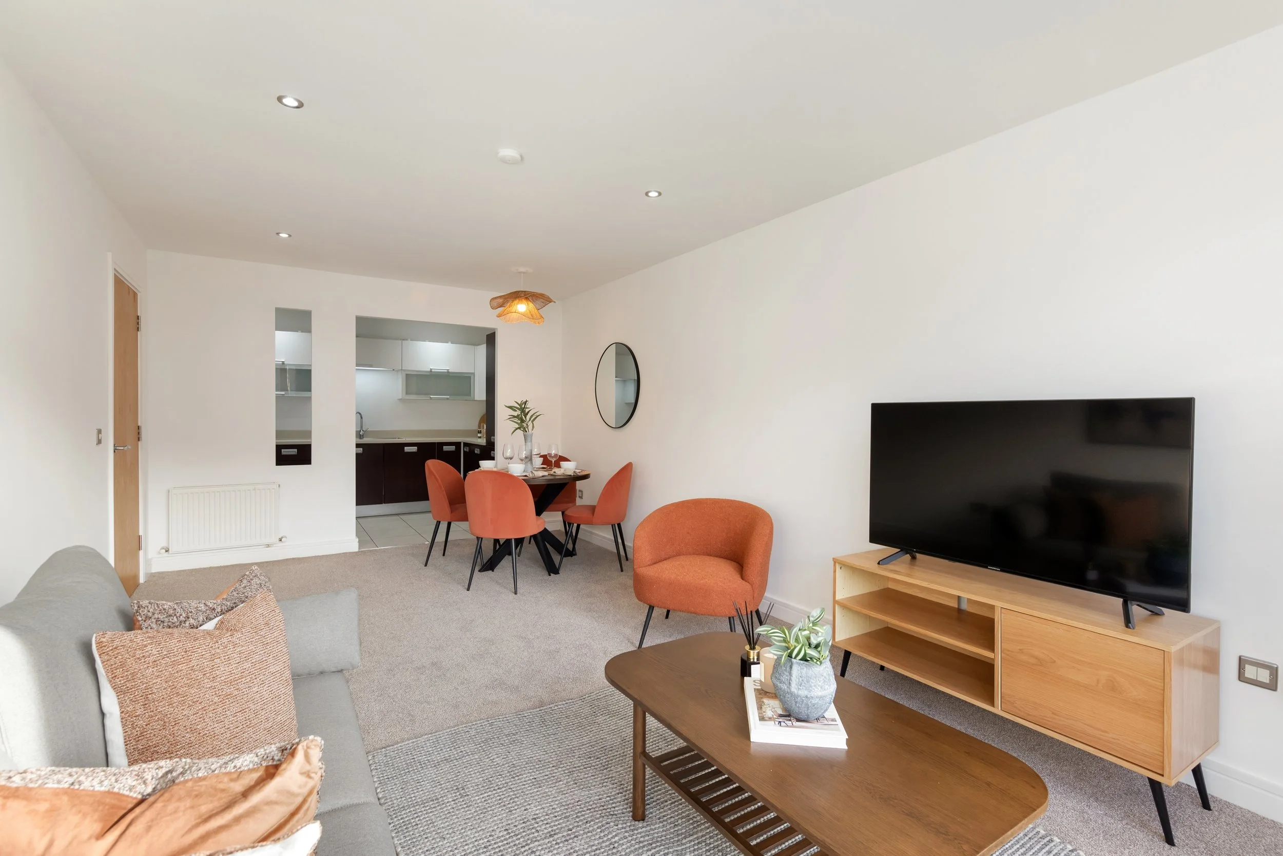 Living room with a gray sofa, orange chairs, a wooden TV stand, and a flat-screen TV, adjacent to a dining area with a round table and four orange chairs, in an open-concept space.