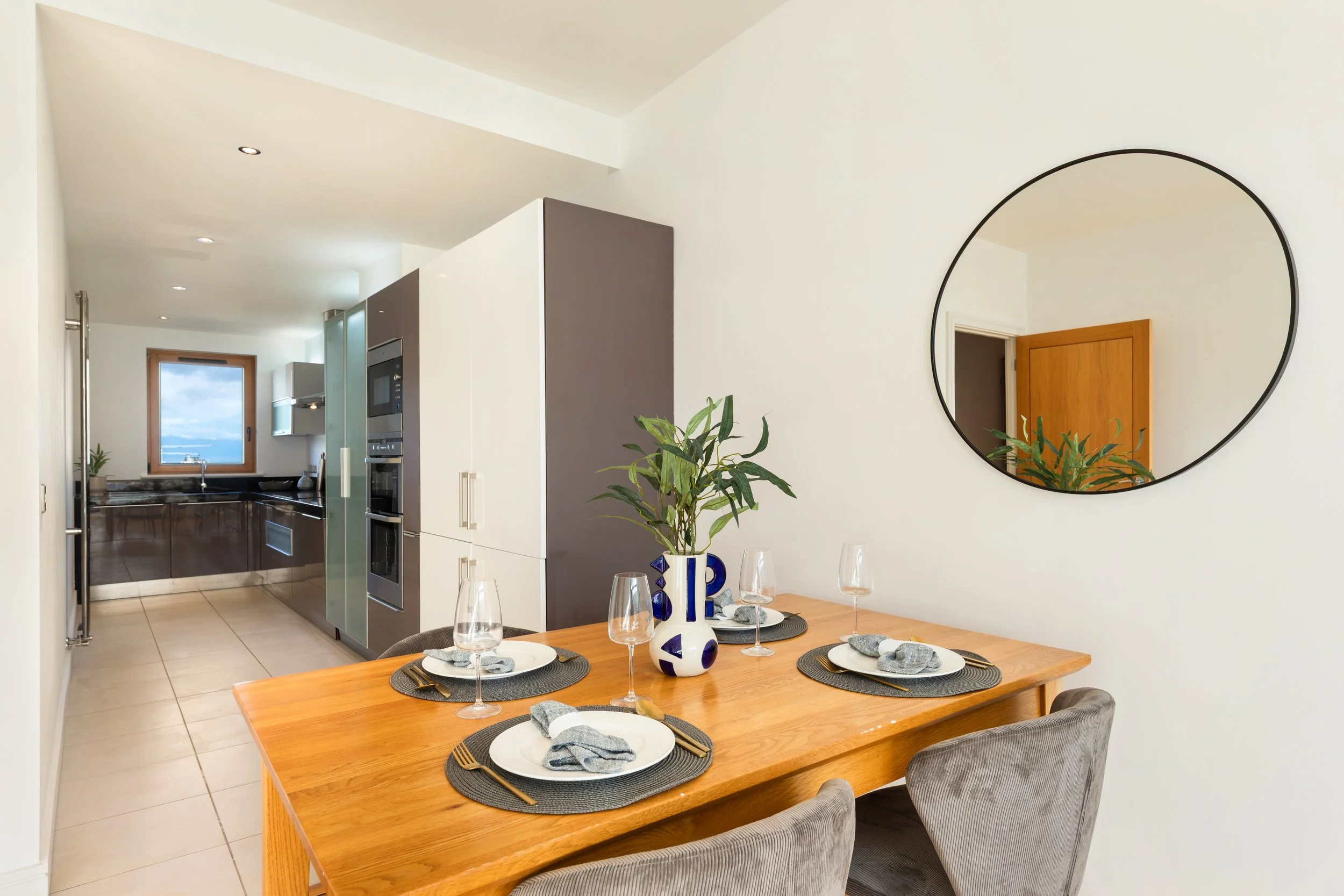 Kitchen and dining area with a wooden dining table set for four, a modern kitchen with white and dark cabinetry, a potted plant, a round mirror on the wall, and a window showing a cloudy sky.