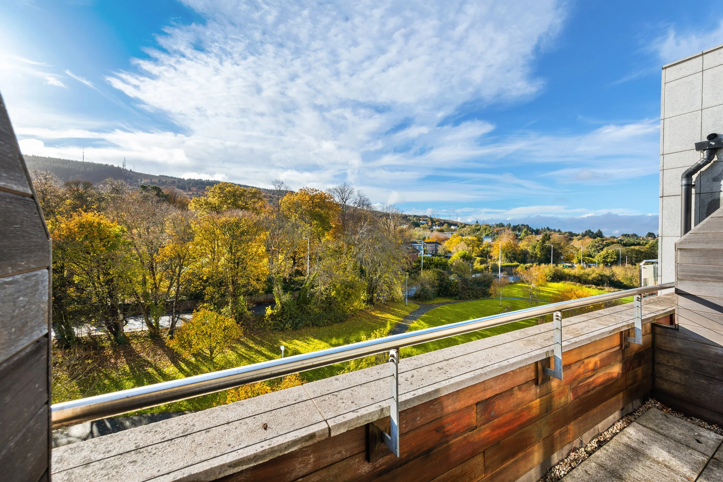 Balcony with a metal railing overlooking a landscape of trees with autumn foliage, grassy areas, and distant hills under a partly cloudy sky.