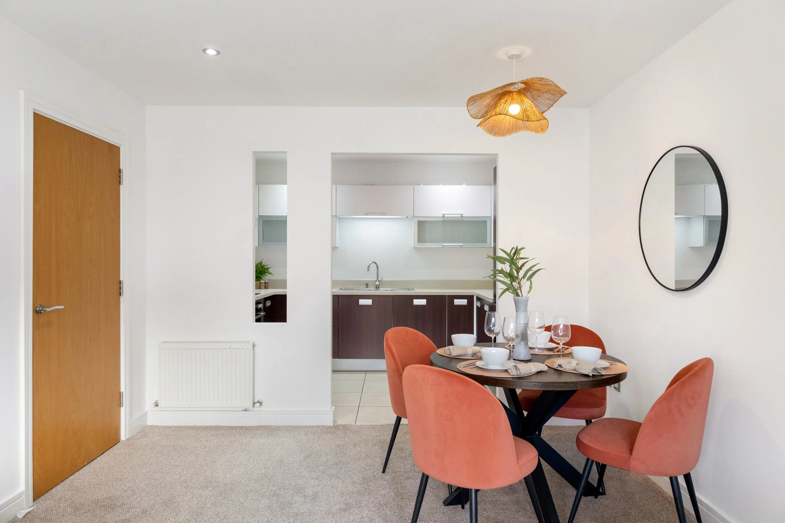 Dining area with four peach-colored chairs around a round dark wood table set with white bowls, glasses, and a potted plant, adjacent to a kitchen with dark lower cabinets and white upper cabinets, a mirror on the wall, and a wooden door