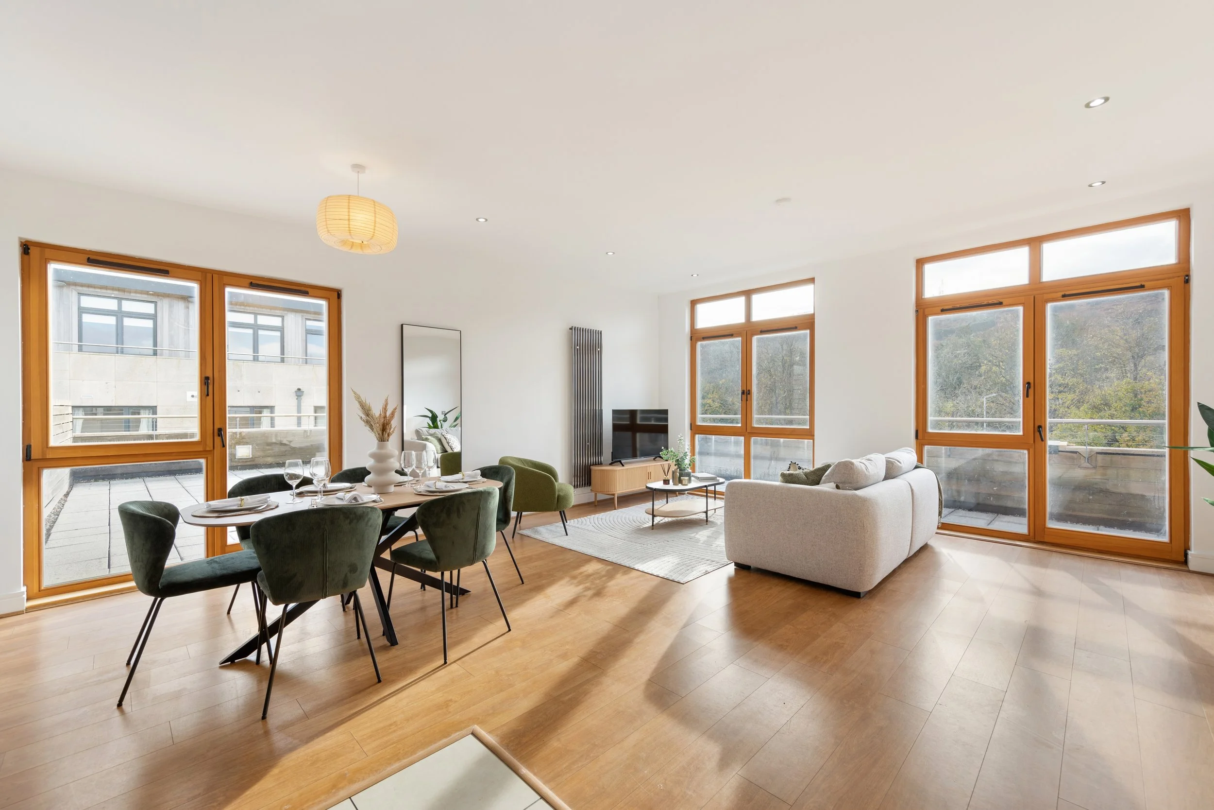 Spacious living and dining area with large windows, wooden floors, white walls, a dining table with six dark green chairs, a white couch, a white rug, a TV on a light wood stand, a tall mirror, and minimalistic decor.