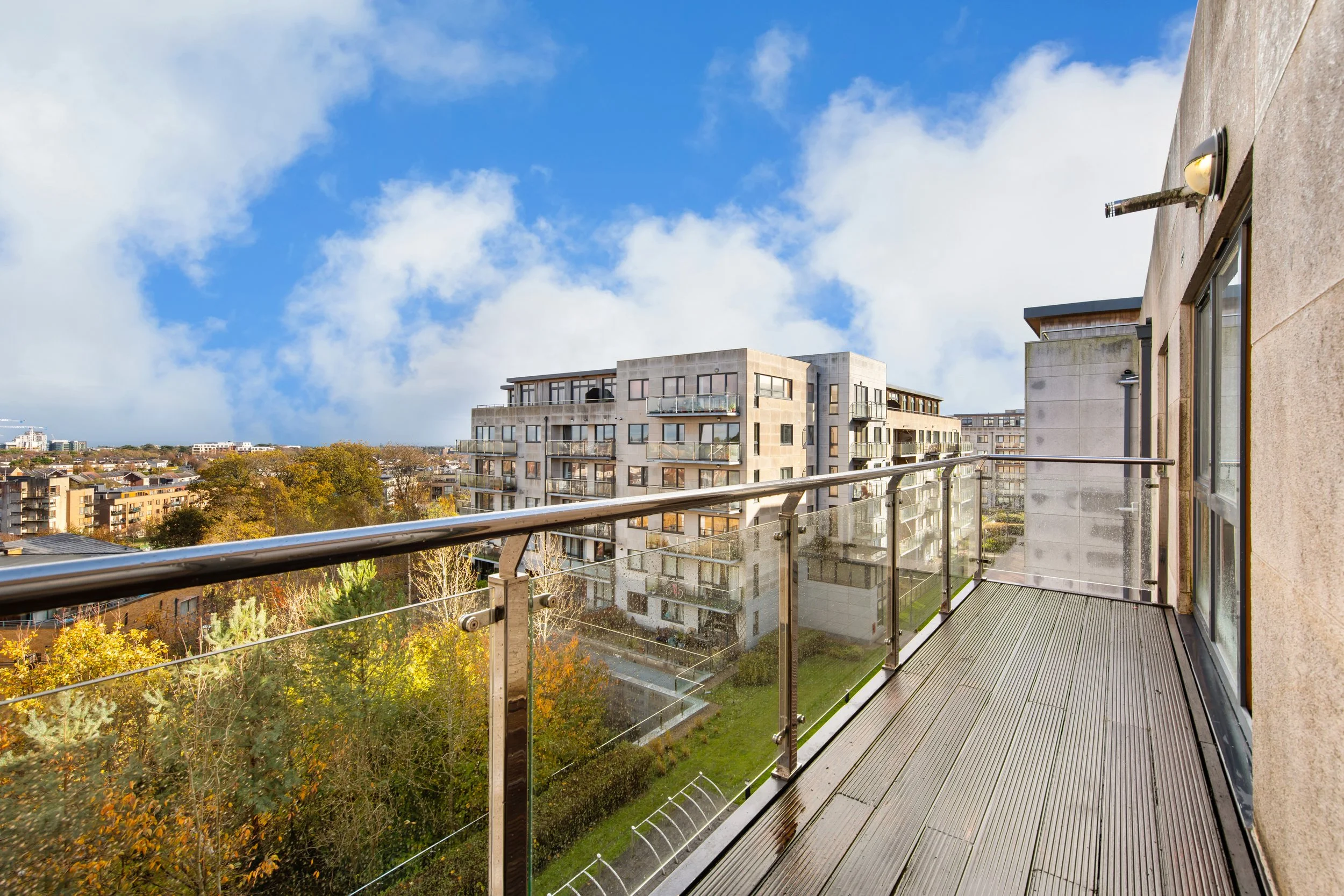 View from a balcony with glass railing overlooking a cityscape with modern apartment buildings, trees with autumn foliage, and a partly cloudy sky.