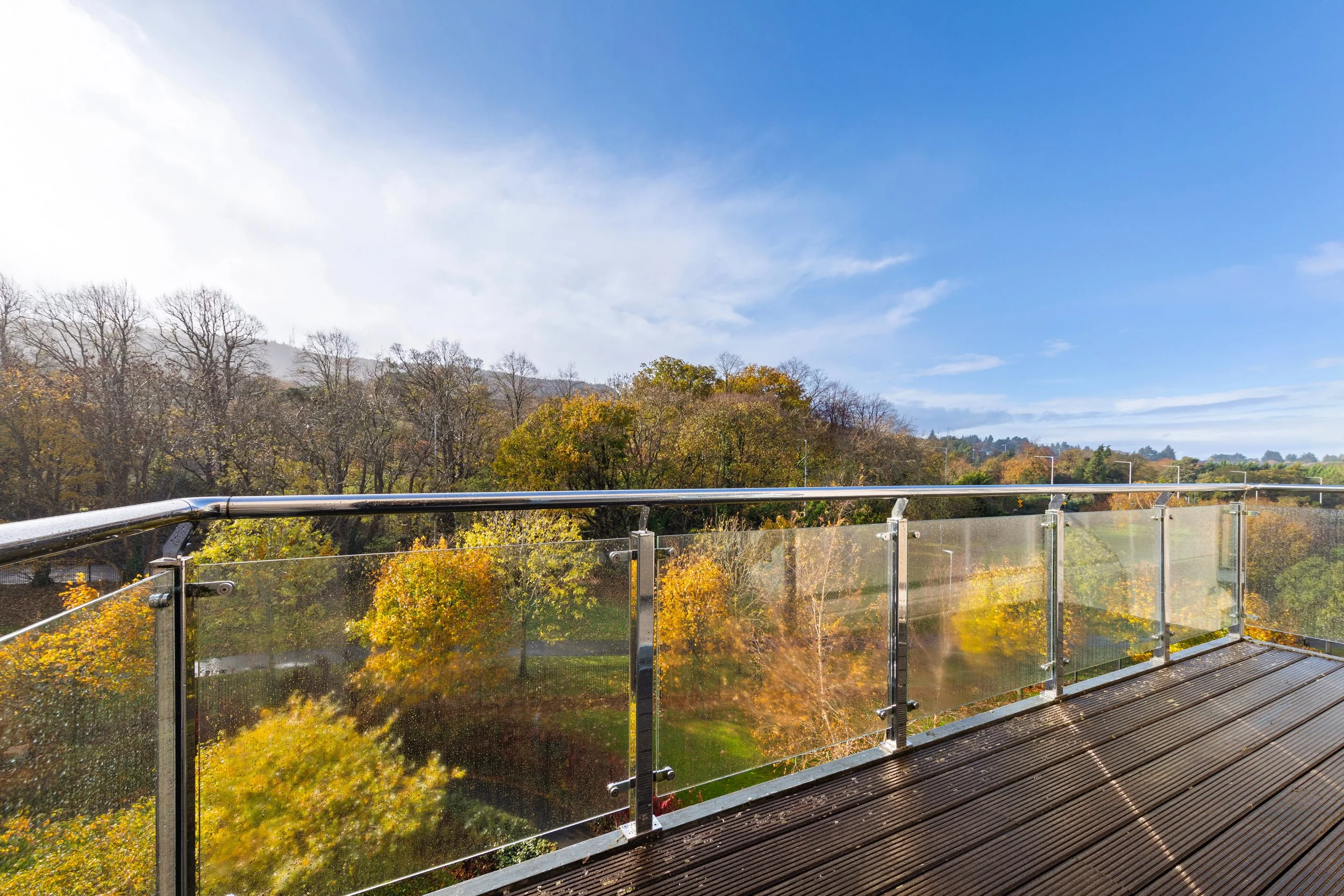 View from a balcony with a glass railing, overlooking a park with trees in autumn colors, under a partly cloudy blue sky.