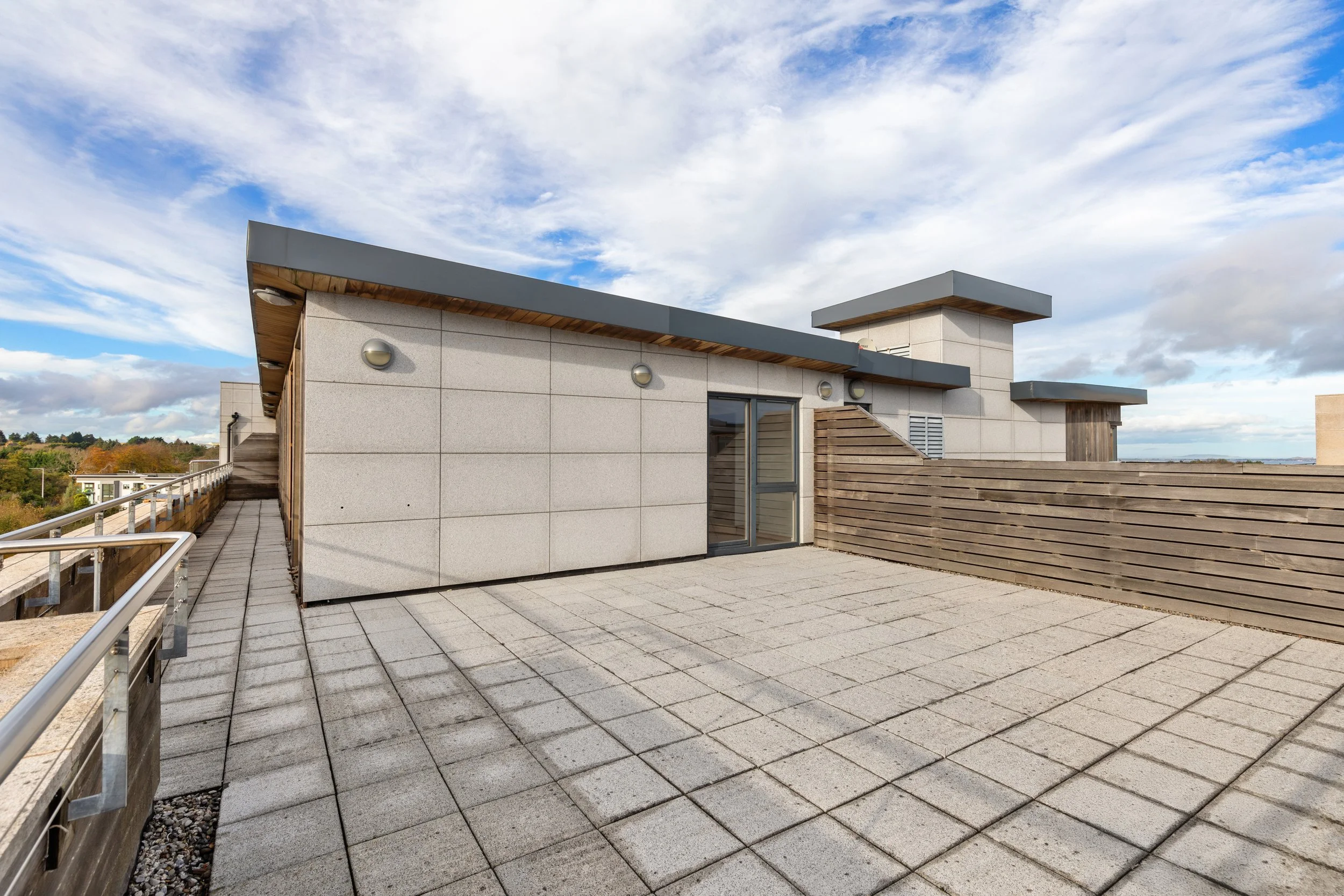 Rooftop terrace with tiled floor, wooden fencing, exterior walls with modern finish, and a glass door under a cloudy sky.