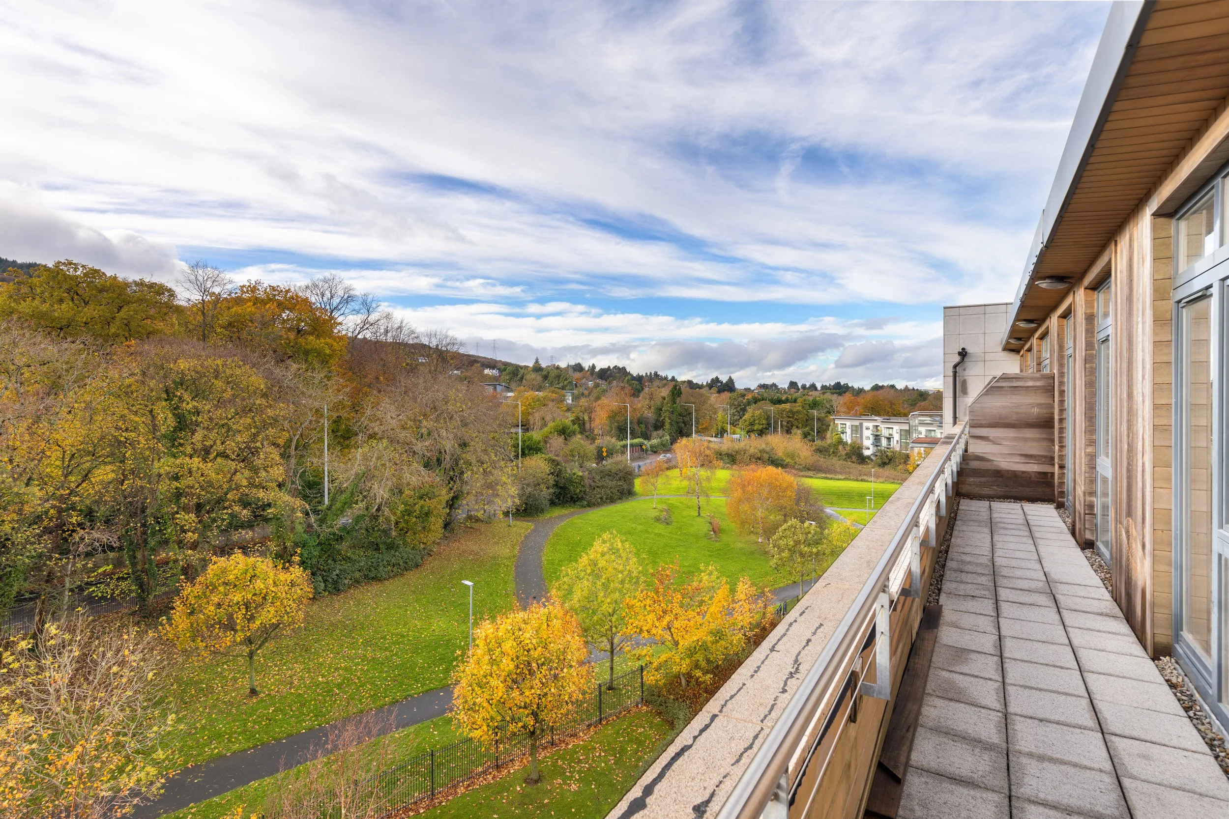View from a balcony showing a green park, trees with autumn foliage, walking paths, and a partly cloudy sky in the distance.