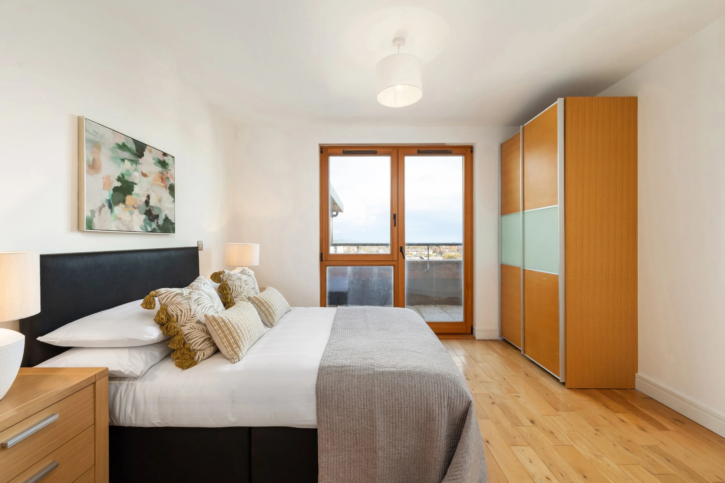 Bedroom with wooden floor, bed with white linens and decorative pillows, two lamps, large window with balcony, colorful abstract wall art, wooden wardrobe.