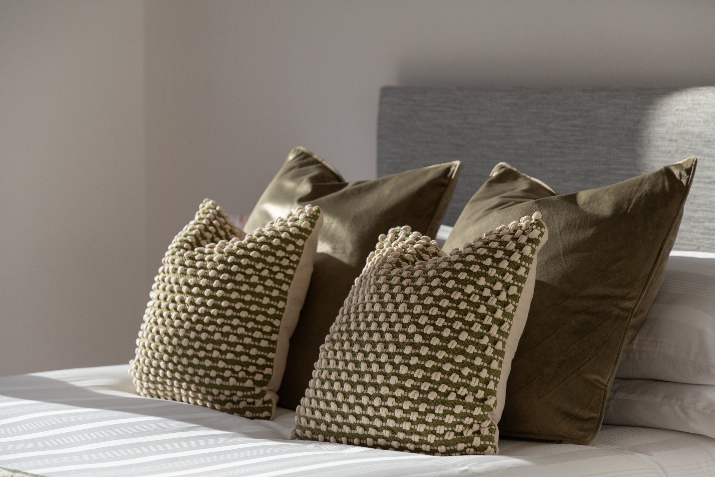 Stack of decorative pillows on a bed, including two textured beige pillows with a knotted pattern and two smooth brown pillows, in a bedroom with a grey headboard.