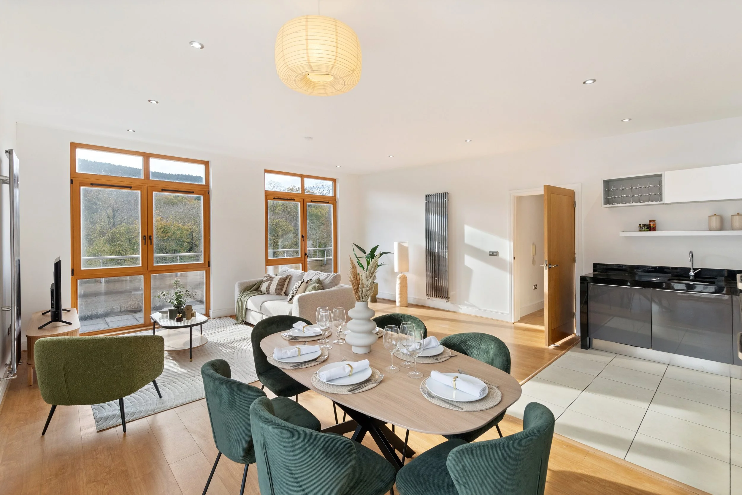 Open-concept living and dining area with large windows, a white sofa, a round dining table set with plates, glassware, and napkins, a small coffee table, and a modern kitchen area featuring black cabinetry and a sink.
