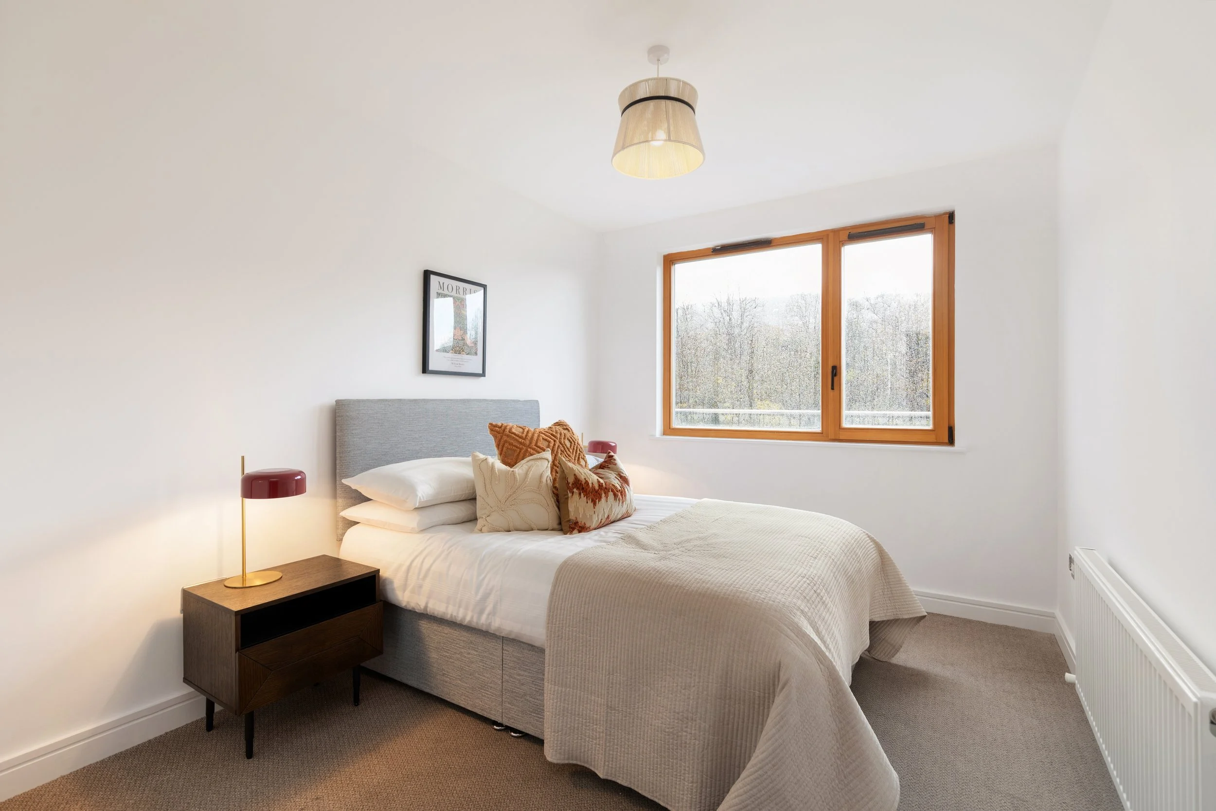 A modern bedroom with white walls, a bed with white bedding and multiple pillows, a wooden nightstand with a red table lamp, a large window showing an outdoor view, and a ceiling light with a beige lampshade.